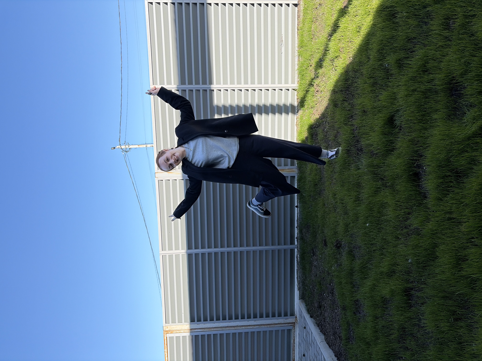 A woman in a black suit and gray sweater is suspended in mid-air, arms outstretched, smiling at the camera. She is wearing black and white sneakers. Behind her is a gray, corrugated metal fence, with a strip of green grass to its right. Above is a clear blue sky, and in the distance, power lines are visible. The image was likely taken outdoors, at a location in the city of Chaikovsky, Russia, characterized by an industrial-looking fence and well-manicured grass. The woman's pose indicates she is likely jumping or being lifted into the air for the photograph.