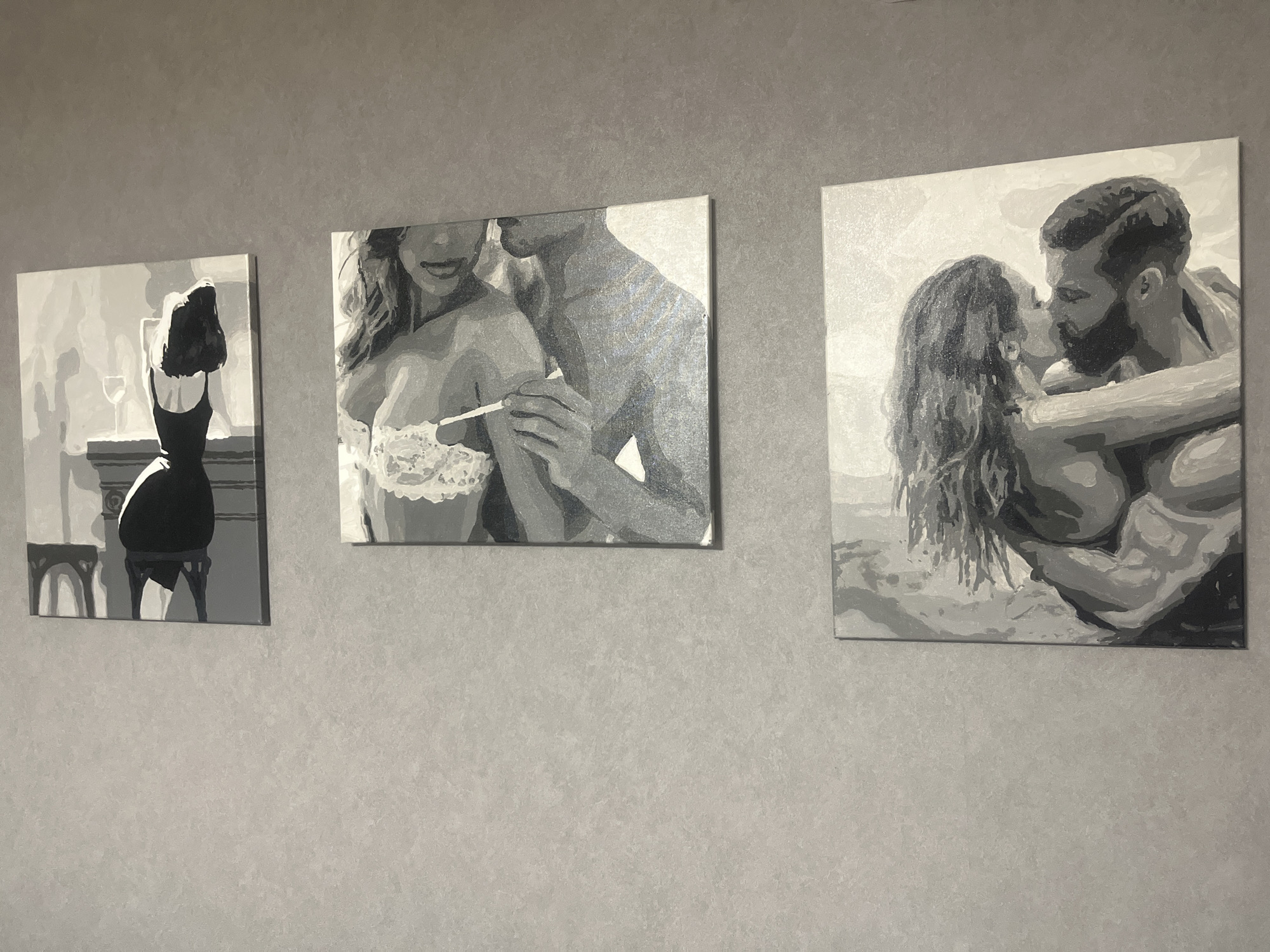 The image shows three black and white paintings hanging on a grey textured wall. The paintings depict romantic scenes between couples. The leftmost painting shows a woman in a black dress viewed from behind, with a wine glass on a table. The middle painting shows a couple embracing, with the woman in a lace bra and the man holding her hand. The rightmost painting depicts a couple kissing intimately. The scene appears to be an interior, likely a home, in the city of Rybnitsa, Moldova.