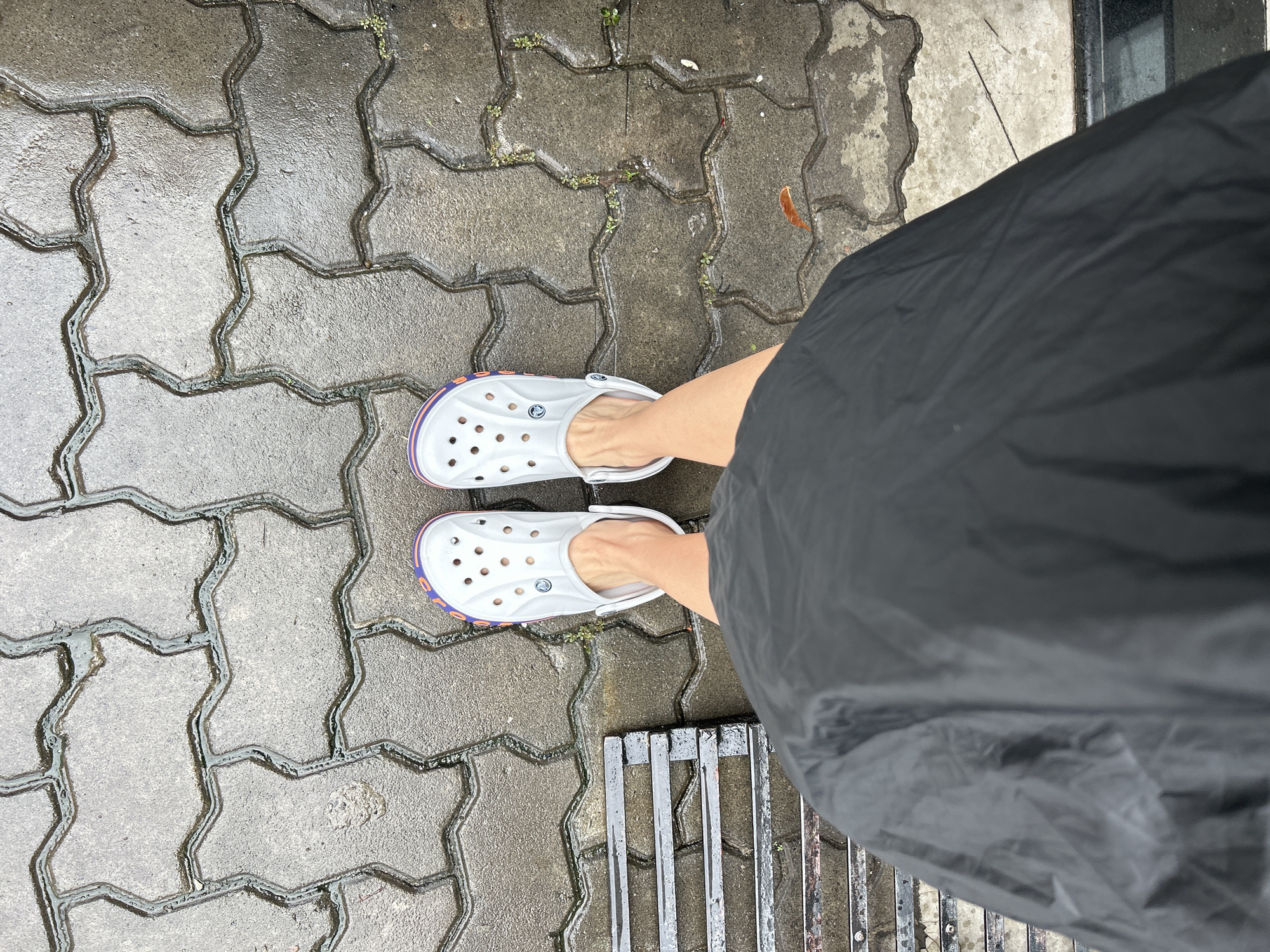 A top-down shot captures a person's feet and lower legs standing on a wet, paved surface. The individual wears light grey Crocs clogs, featuring purple and orange trim along the sole, with the Crocs brand logo visible on the upper part of each shoe. Bare feet and ankles are visible within the clogs. A dark, possibly black, garment, likely a skirt or rain gear, extends from the right side of the frame, covering most of the body below the knees.
The ground consists of interlocking grey concrete pavers, which are visibly wet, indicating recent rain. Small clusters of green weeds grow in the cracks between the pavers, and a few dried brown leaves are scattered on the wet pavement. A metal drainage grate is partially visible in the bottom right corner of the frame. The scene suggests an outdoor environment during or after rainfall, set in a location like Kochi, India.