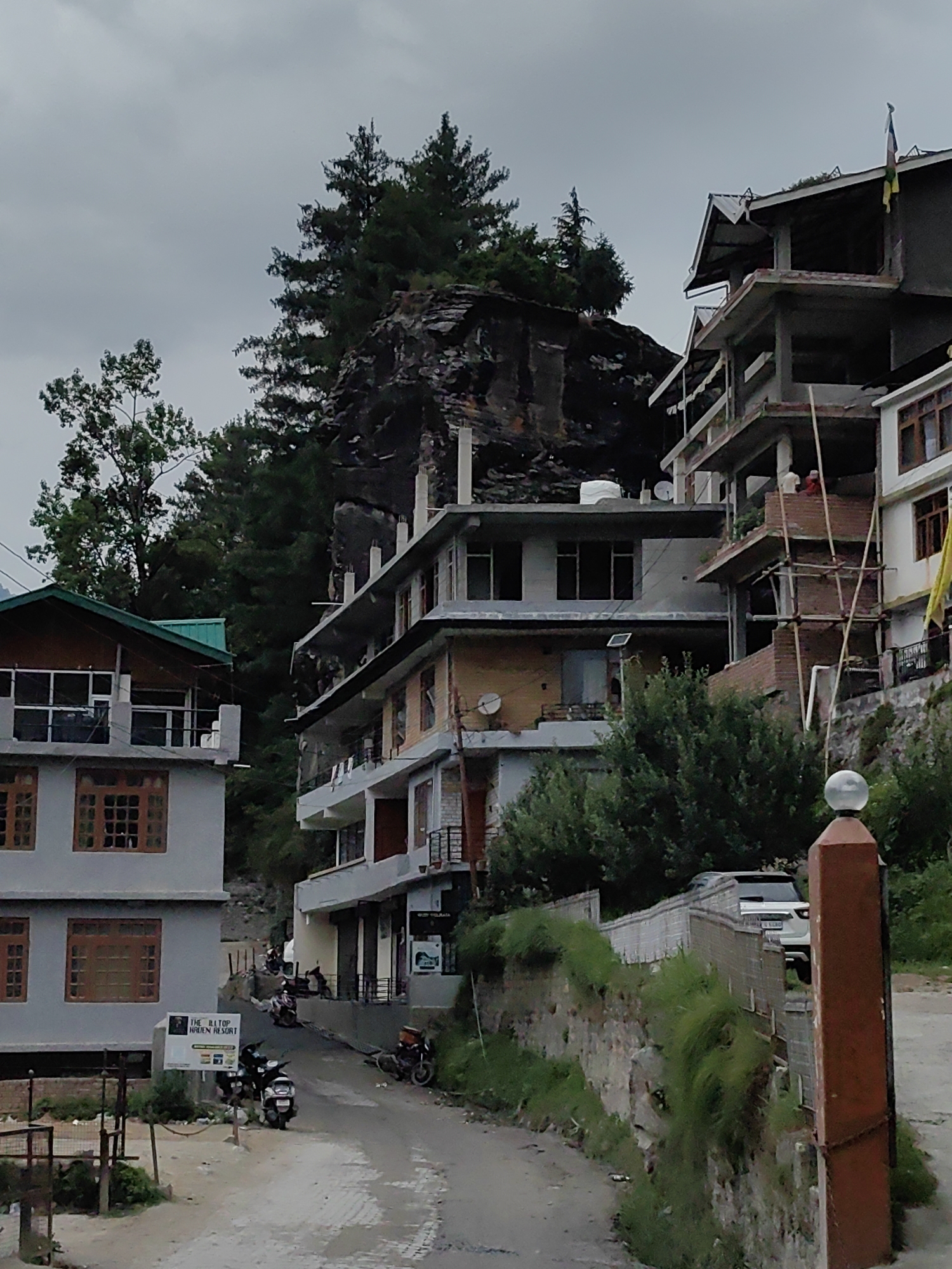 The image depicts a street scene in Manali, India, characterized by a narrow, paved road winding uphill between buildings. The architecture is a mix of modern and under-construction structures, with several multi-story buildings featuring balconies and exposed brickwork. Vegetation, including tall pine trees and lush green bushes, is interspersed with the buildings, suggesting a mountainous or hilly terrain. The sky is overcast with a dull, cloudy appearance.
In the foreground, to the left, is a grey building with wooden-framed windows and a green-roofed section. A sign for "THE HILLTOP HAVEN RESORT" is visible on this building. Several motorcycles and scooters are parked or being ridden on the road. To the right, a stone wall with overgrown grass separates the road from a higher elevation where another building is under construction, with scaffolding and apparent workers. A white SUV is parked on the higher level. A reddish-brown pillar with a glass fixture stands prominently in the right foreground. The overall impression is one of a developing tourist destination in a natural, hilly environment.