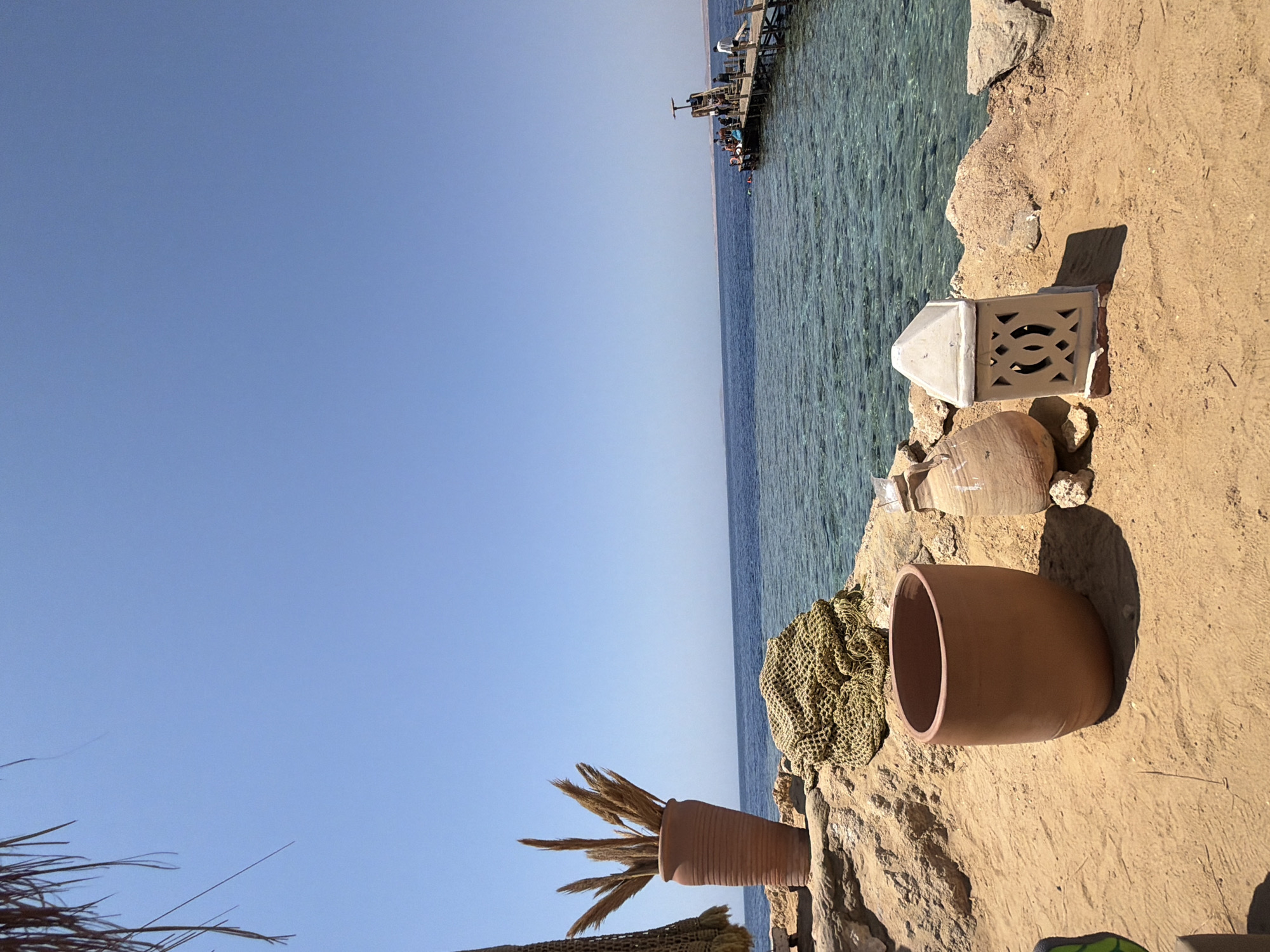 This image depicts a serene coastal scene at the Red Sea in Sharm El Sheikh, Egypt. In the foreground, on a sandy area next to rocky outcrops, are several decorative objects. A large, terracotta vase filled with dried reeds stands prominently. Next to it, a pile of coarse, woven netting rests on the rocks. Further to the right, another, wider terracotta pot is visible, and to its right, a decorative lantern with intricate geometric cutouts. In the background, the clear blue waters of the Red Sea stretch to the horizon under a clear sky. A wooden pier extends into the sea, with several people visible on it and in the water, suggesting a popular spot for swimming or water activities. The overall impression is one of a tranquil, sunny day in a popular tourist destination.