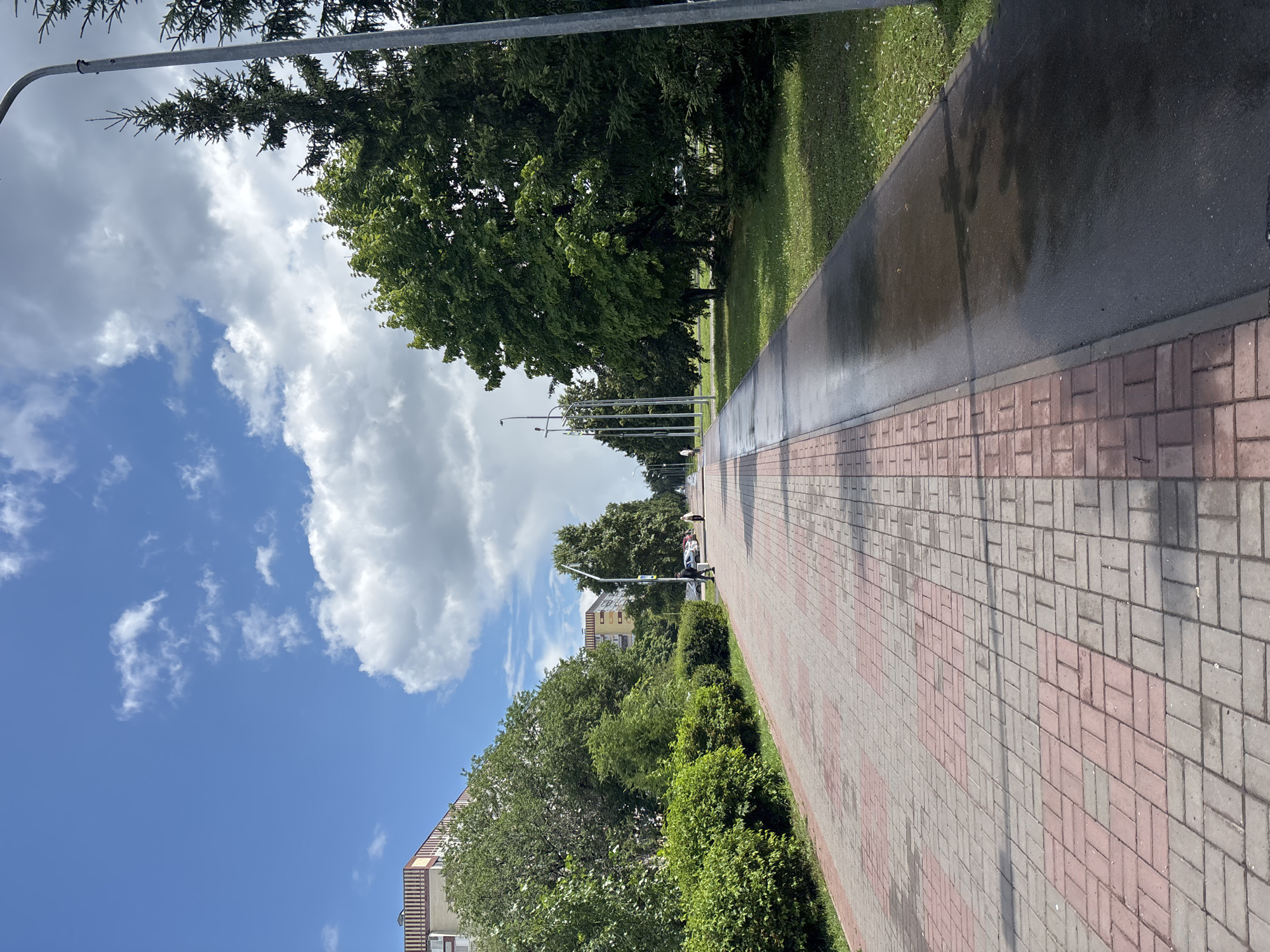 A paved walkway with alternating red and gray bricks leads into the distance, bordered by a wet, dark asphalt path on the right and a row of green bushes on the left. Beyond the bushes, several trees and buildings are visible. Further down the walkway, two individuals are walking. The sky above is a vibrant blue, dotted with fluffy white clouds, and trees line the right side of the path, with a tall lamppost visible. The scene appears to be a residential area in Naberezhnye Chelny, Russia, likely a park or a tree-lined street.
