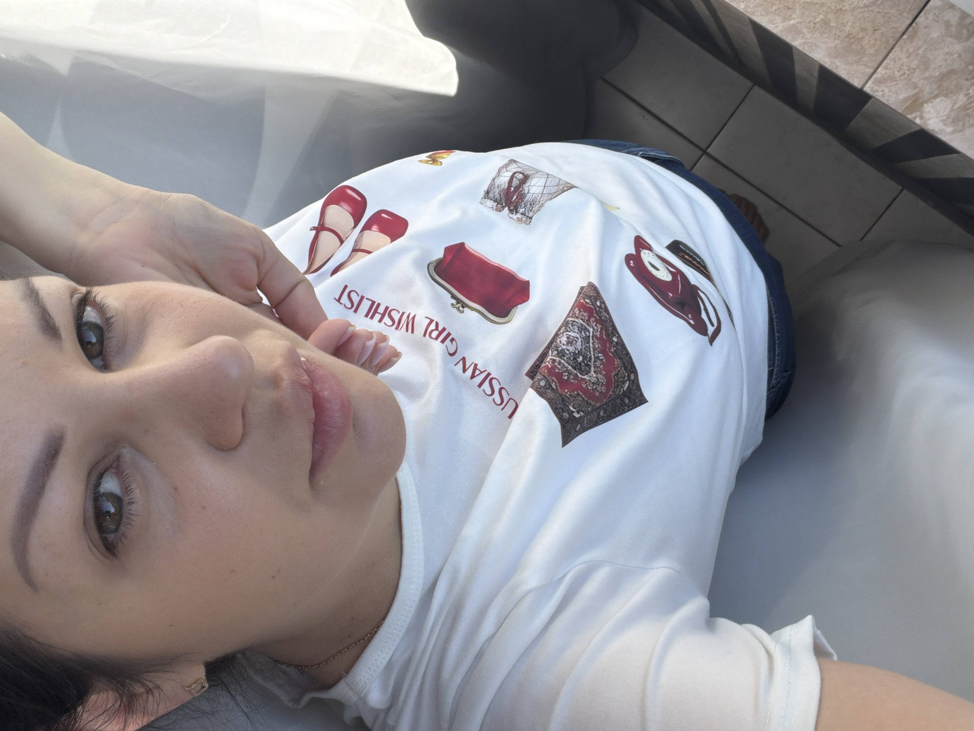 In Sochi, Russia, a woman with dark hair and green eyes poses for a selfie, her face filling the top portion of the frame. She wears a white t-shirt adorned with various illustrations, including red ballet flats, a red clutch purse, and a small Persian rug. Her right hand is raised, touching her face. The background is a light gray wall and a window with sheer white curtains that cast a soft light. The lower portion of the image shows her torso and a hint of blue jeans. The scene appears to be indoors, with tiled flooring visible at the bottom.