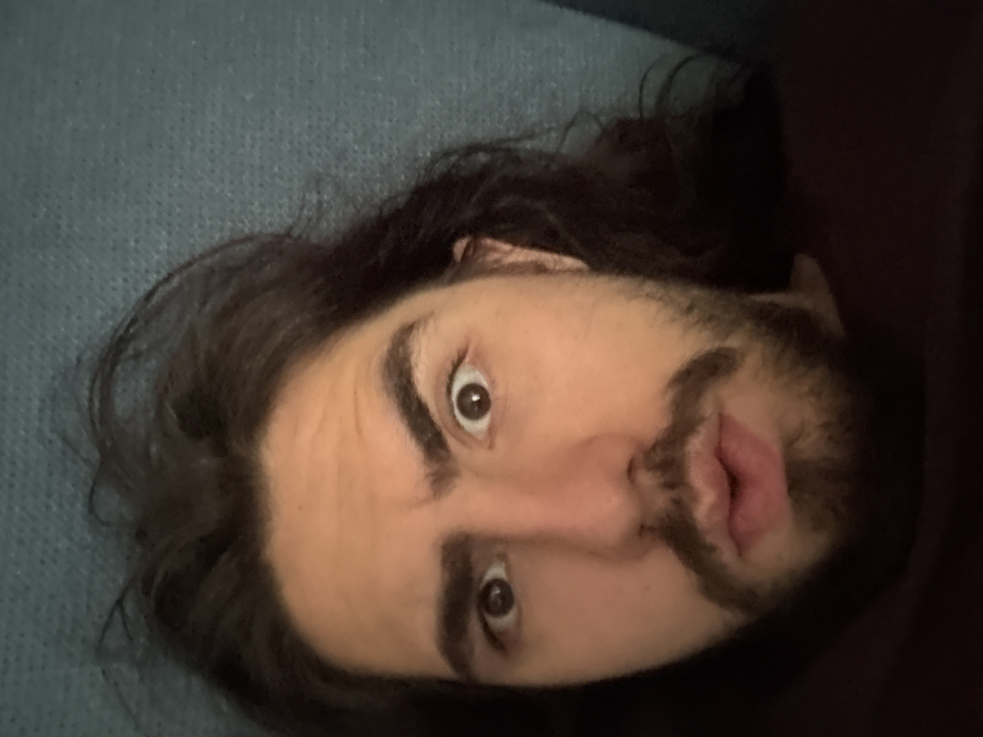 A close-up, high-angle selfie shows the face of a person with dark hair and a beard, looking directly at the camera with wide eyes and a slightly open mouth. The person has thick, dark eyebrows and dark brown eyes. Their hair is long and appears somewhat disheveled, framing their face. The background is a textured, dark teal surface, likely a wall or fabric. The lighting is dim, casting soft shadows on the person's face. The overall impression is one of surprise or curiosity.