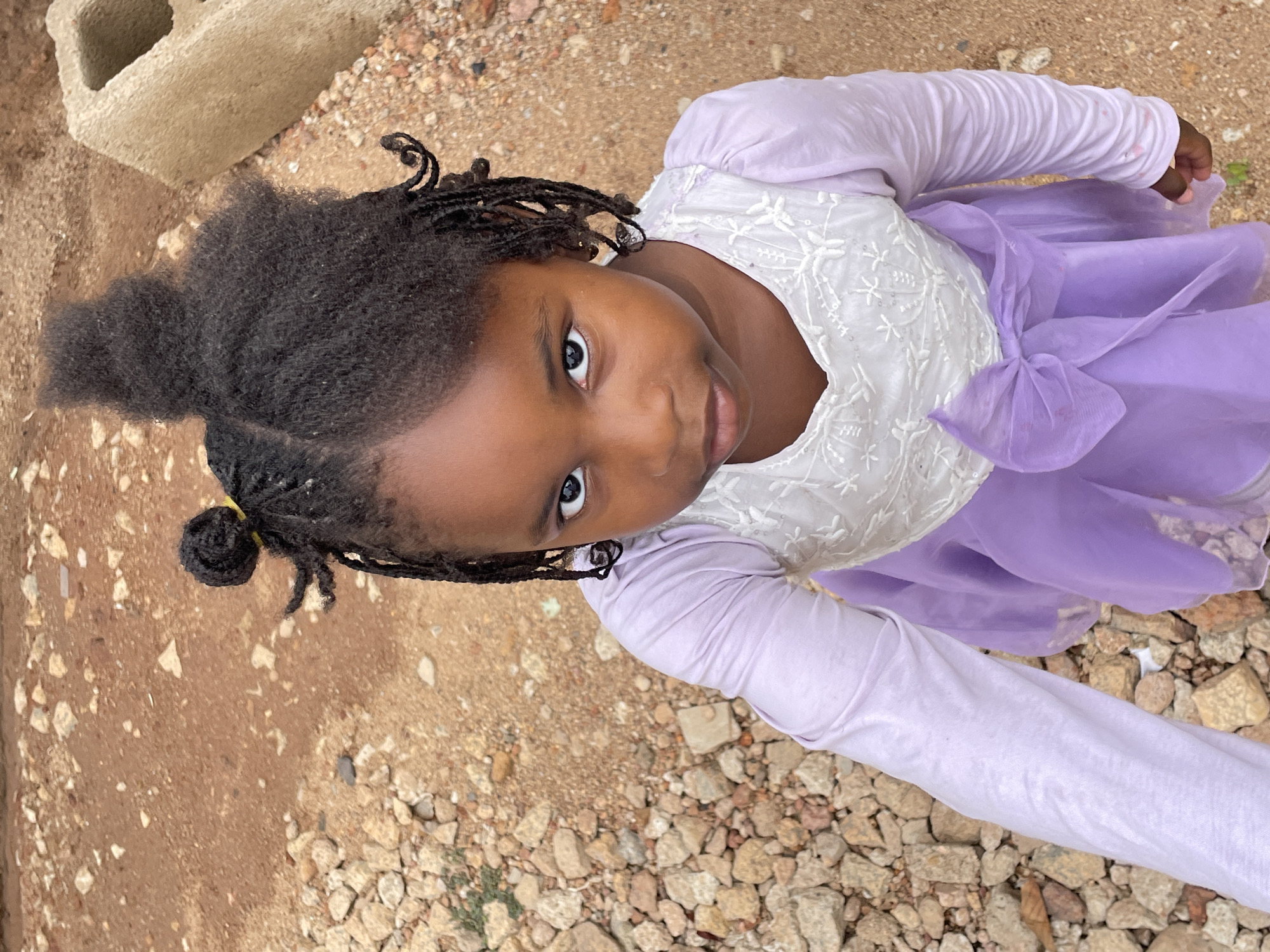 A young girl stands in an outdoor environment in Jos, Nigeria, looking up at the camera. She is wearing a light purple dress with a white, lace-detailed bodice and long sleeves. Her hair is styled with twists and a section of loose hair on top. The ground beneath her feet is a mixture of red soil, gravel, and small rocks. A grey concrete block is visible in the upper left corner of the frame.