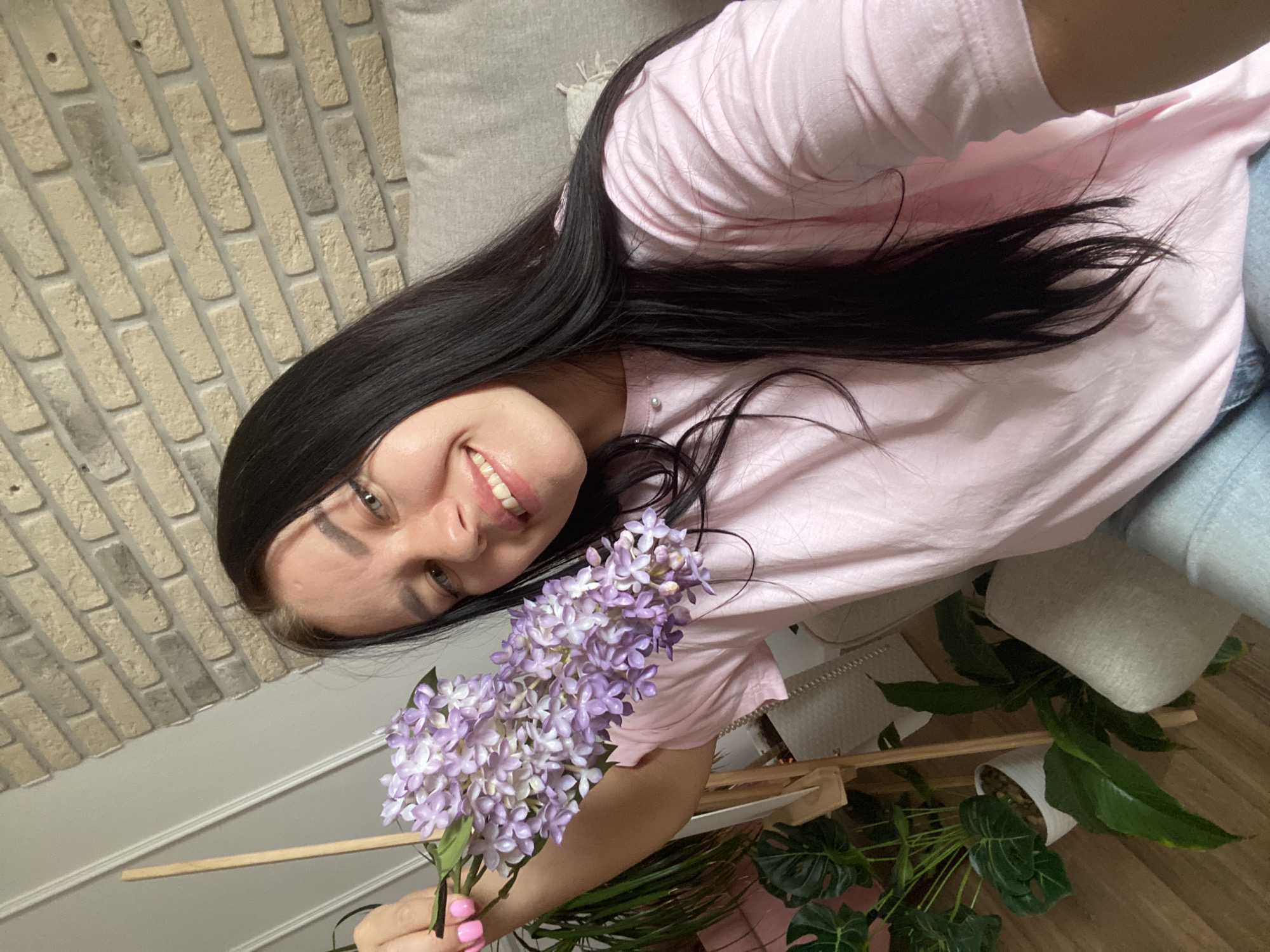 A young woman with long, dark hair and pale skin is holding a sprig of lilac flowers in her left hand. She is smiling, revealing white teeth, and her eyes are open. She is wearing a light pink t-shirt. The background includes a light-colored brick wall and a beige sofa. Several potted plants are visible to the left of the woman. The scene appears to be indoors, likely in a residential setting, and based on the available information, the location is the city of Chaikovsky, Russia.