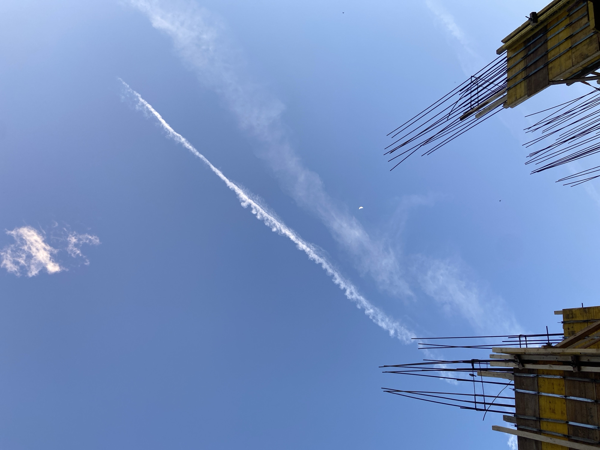 The image shows a bright blue sky with a diagonal contrail and some scattered clouds. In the bottom right and upper right corners, there are structural elements with protruding rebar, suggesting a construction site. A small object, possibly a bird or an aircraft, is visible near the center of the frame. The location is presumed to be a construction site in an urban area, city and state unknown.
