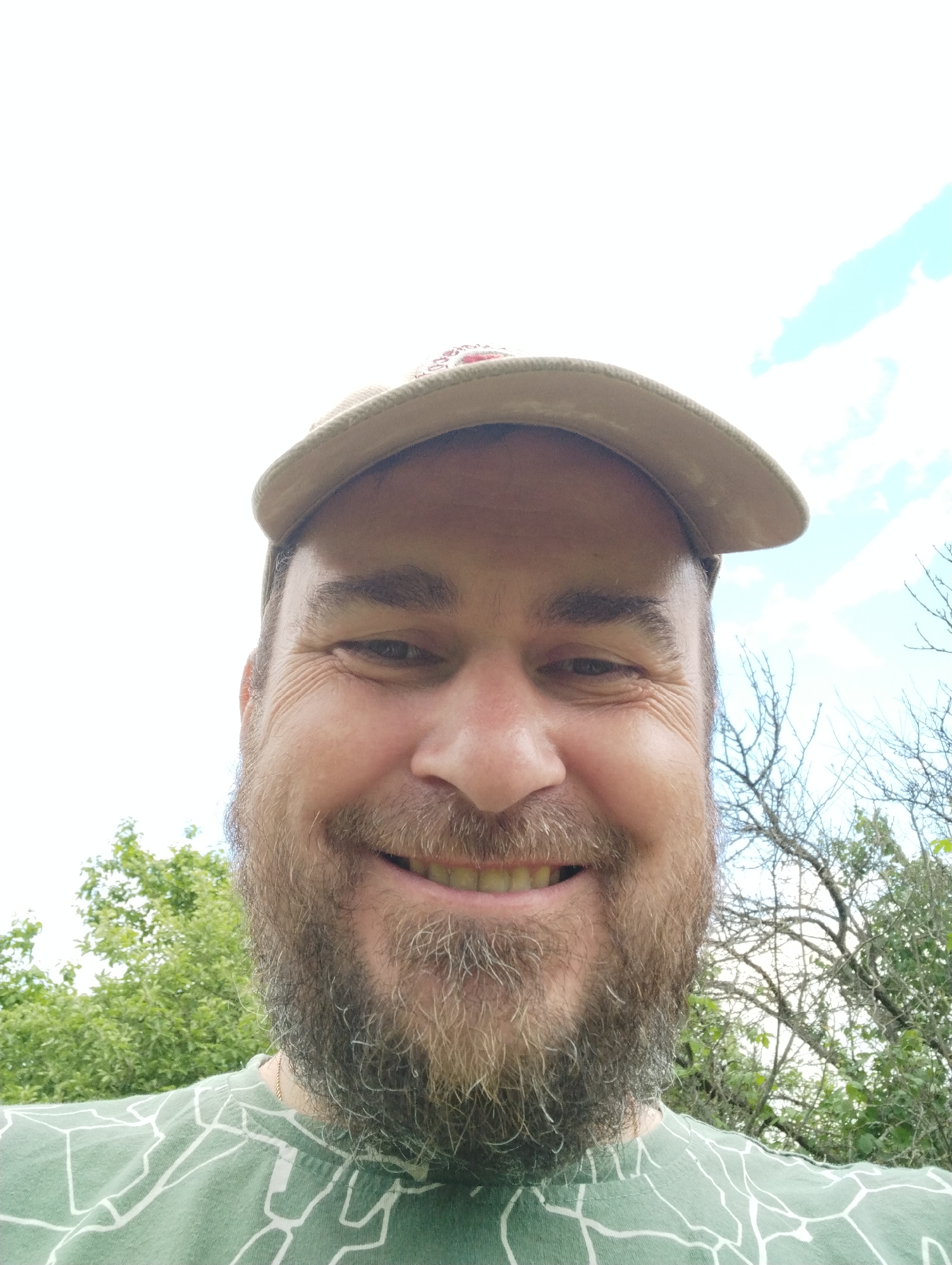 This media file is a selfie of a man in his 40s, taken outdoors. He is wearing a light-colored baseball cap and a green t-shirt with a white abstract pattern. He has a full beard and is smiling, showing his teeth. His expression suggests a cheerful or friendly demeanor.
The background consists of lush green trees, some of which have bare branches, suggesting it might be late spring or early autumn. The sky is bright and appears to have some light clouds, indicating daytime. The overall setting is natural and rural, consistent with the provided location context of Ширяево (ильинский), Russia, a known natural park and village. There is no visible text in the image. The selfie is taken from a slightly low angle, looking up at the man.