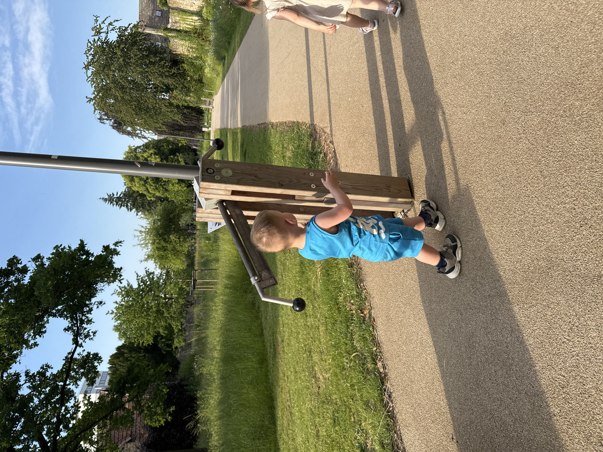 In the city of Unknown, Unknown, a young boy wearing a blue sleeveless shirt and blue shorts walks on a paved path next to a wooden structure with metal exercise equipment. He is looking towards the right side of the image, where a young girl in a light-colored dress and sandals is also walking away from the camera on the same path. The path is bordered by a grassy area with tall grass and trees. In the background, there is a stone wall covered in ivy, more trees, and buildings. The sun is casting long shadows across the path, indicating it is late afternoon or early evening.