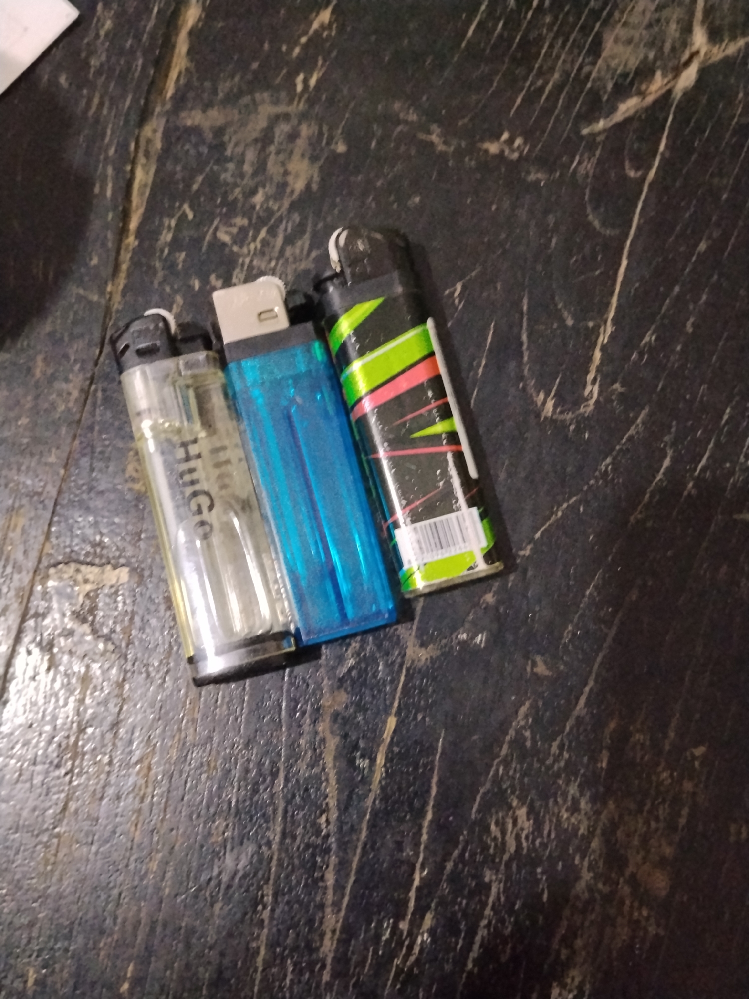 Three lighters are arranged on a dark, textured wooden surface, possibly a table. From left to right, the first lighter is clear with a visible liquid fuel inside and bears the brand name "Hugo" printed vertically in black. The second lighter is a translucent bright blue. The third lighter is black with a design featuring bright green and pink diagonal lines and a white barcode sticker at its base. The wooden surface exhibits significant wear, with visible scratches and grain patterns. The lighting is dim, creating shadows and highlighting the textures of both the lighters and the surface. The location in the city of Bilis Bilis, Indonesia, is not discernable from the image.