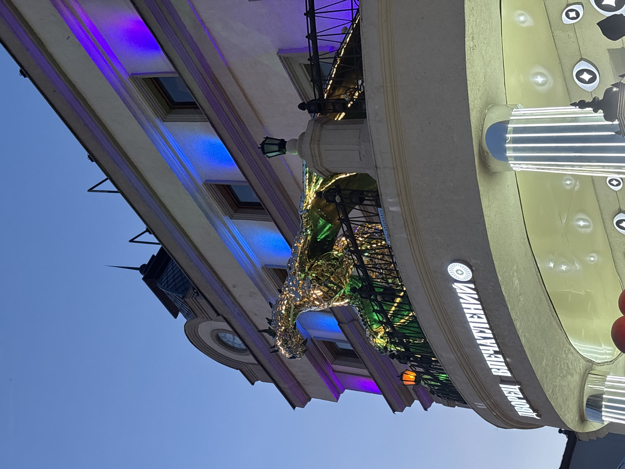 The image shows the exterior of a building in Kazan, Russia, featuring a large, metallic dragon sculpture on a balcony. The building is illuminated with purple and blue lights. Below the balcony, a sign in white lettering reads "ДВОРЕЦ ВПЕЧАТЛЕНИЙ" (Palace of Impressions). The sky is clear and blue, suggesting it is either early morning or late evening. The dragon sculpture appears to be made of a reflective, possibly metallic, material and is detailed with scales and wings. The balcony railing is ornate, with metalwork. In the foreground, there are decorative elements on the building's facade, including vertical light fixtures and circular motifs that resemble eyes. The overall scene depicts an architecturally interesting building with a fantastical element.