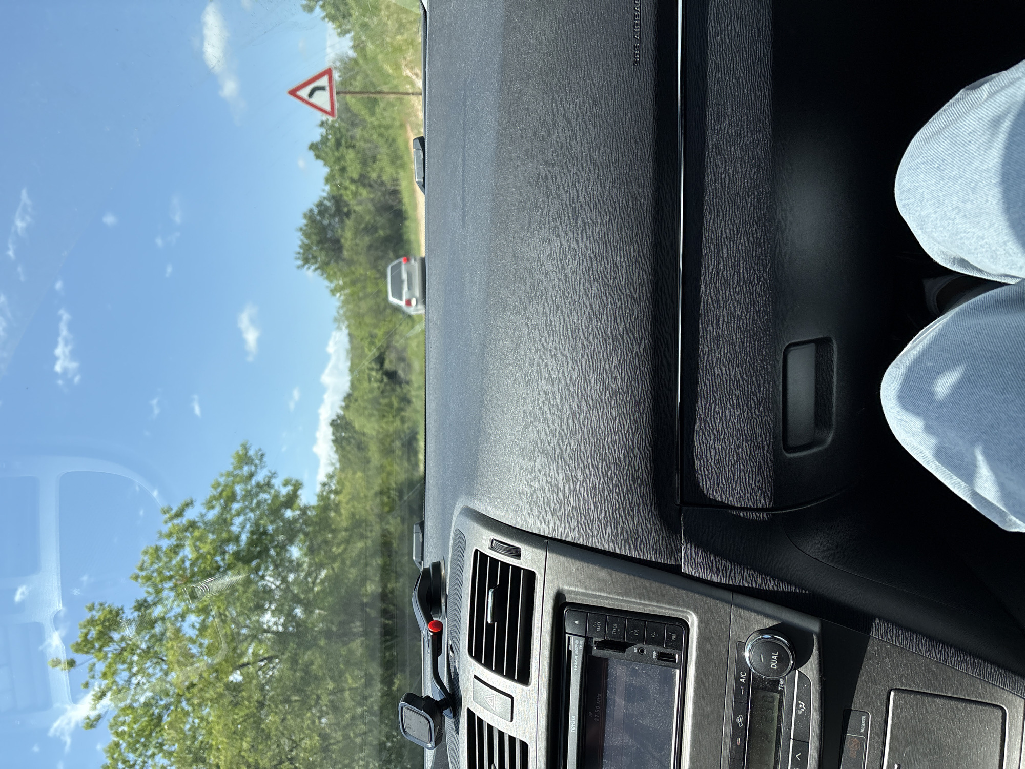 The image is taken from inside a car, focusing on the dashboard and the view through the windshield. The dashboard is black and gray, with visible air vents and a car stereo displaying "87.50 MHz" and buttons for "TRACK," "VOL," "A/C," "TEMP," and "DUAL." The driver's leg is visible on the right, wearing light blue jeans. Outside the car, a road winds through a green, wooded area under a bright blue sky with scattered clouds. A road sign indicating a curve to the right is visible. Another car is ahead on the road. The image provides no explicit information to confirm that the location is in the city of Levski, Bulgaria.