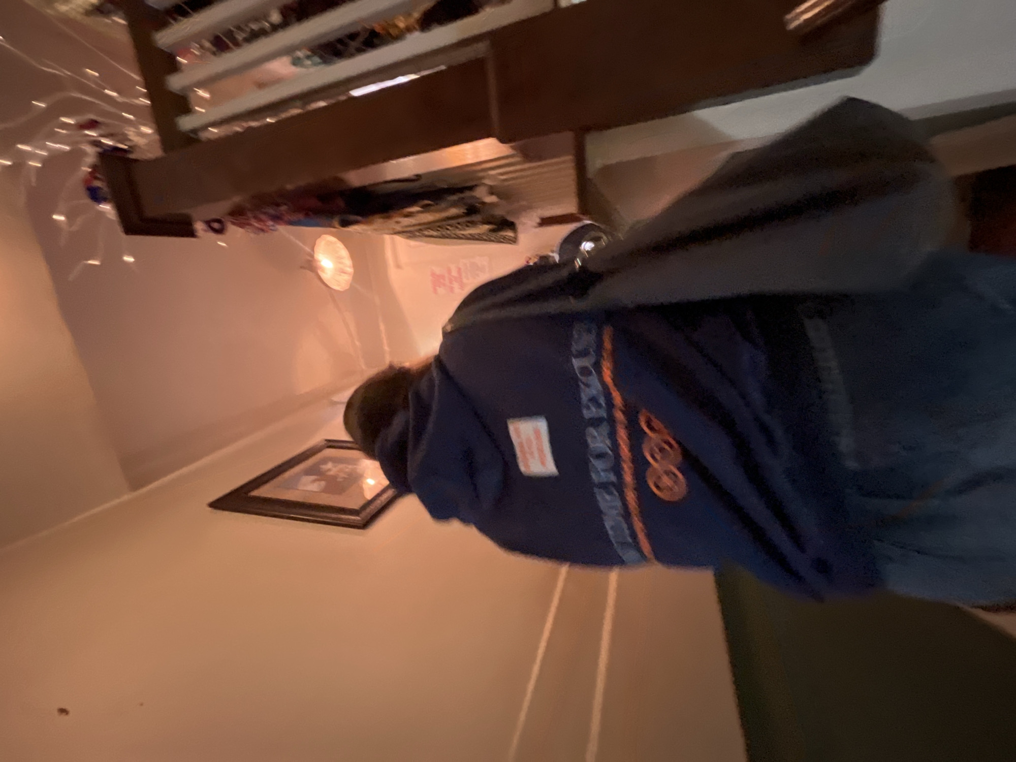 Indoor scene with a person wearing a blue hoodie with orange text and design. The hoodie features a white patch on the upper left area. A framed picture hangs on the wall. Overhead, there is an architectural element with decorative lights. The image was taken indoors. Specific location in New Braunfels, United States of America, cannot be determined from the image.