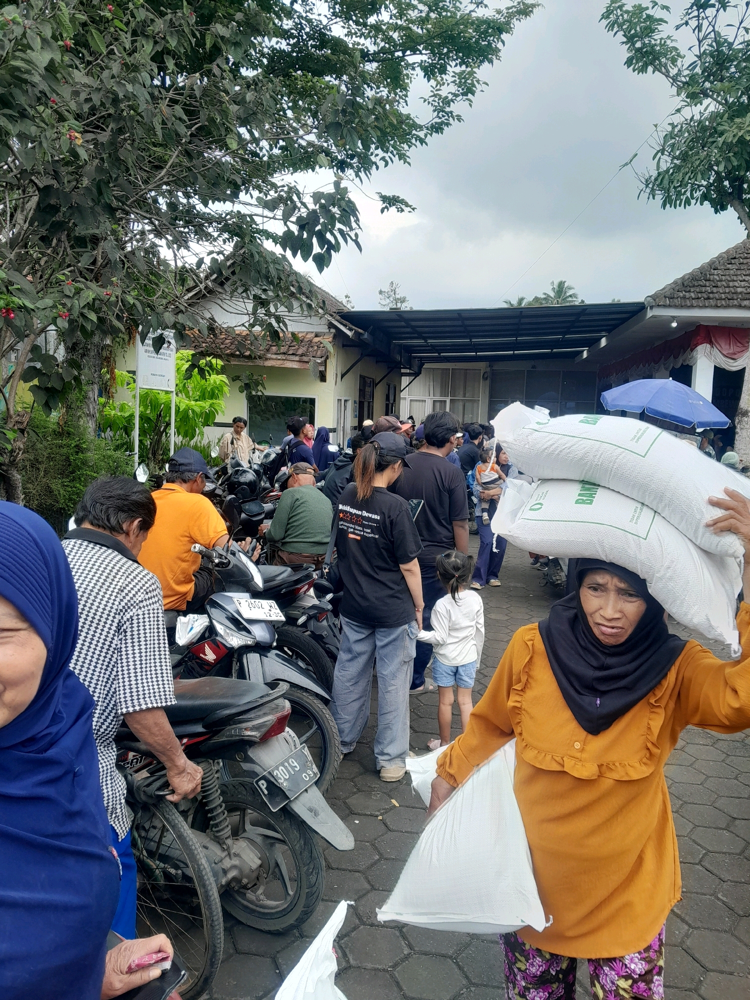 The image depicts an outdoor scene in what appears to be a village or rural setting in Jambewangi, Indonesia. A group of people are gathered around several parked motorcycles, suggesting a distribution or collection point. In the foreground, a woman wearing an orange shirt and a black headscarf is carrying two large sacks of what appears to be rice on her head. She is also holding another sack in her left arm. To her left, another person wearing a blue headscarf is visible. Behind them, a crowd of people, some of whom are seated on motorcycles, are waiting. A signpost with Indonesian text is visible in the background, along with a building and trees. The sky is overcast, indicating a cloudy day. The overall atmosphere suggests a community event or aid distribution.