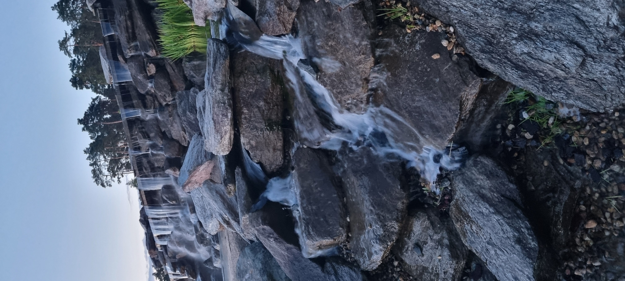 The image captures a man-made tiered waterfall constructed with large, dark gray rocks. The water cascades down the rocks, creating a blurred, silky effect due to a long exposure. Pine trees are visible in the background, lining the top of the waterfall structure. The sky is a pale blue with hints of clouds, suggesting either early morning or late evening. The foreground is dominated by more rocks and pebbles, with some sparse green grass growing amongst them. The scene is likely an outdoor recreational area or park in Shulgino, Russia, designed to mimic a natural landscape with its rock formations and water features. No people or animals are visible in the image.