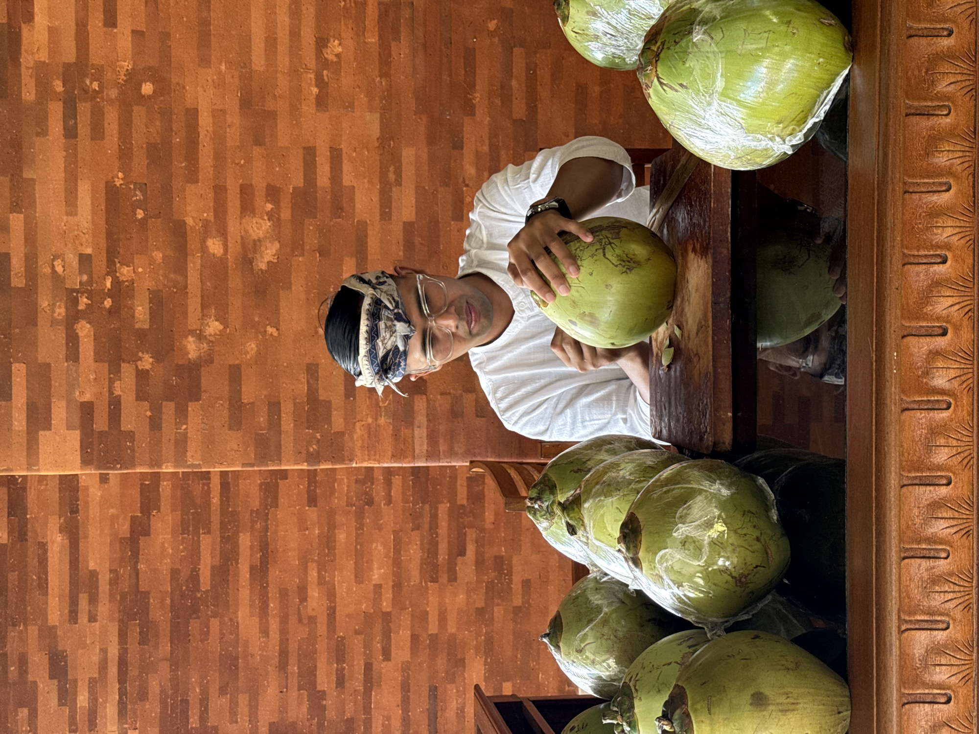 A man, identified as having dark hair and wearing glasses, a white shirt, and a patterned headscarf, is seated at a wooden table in front of a brick wall in Kuta Selatan, Indonesia. He is in the process of preparing a green coconut, holding it in his left hand and using a tool, possibly a knife, with his right hand to peel it. Several other green coconuts, some wrapped in plastic, are arranged on the table in front of him. The scene appears to be indoors, with wooden furniture and a tiled floor reflecting the objects on the table. The brick wall behind the man features a repeating horizontal pattern.