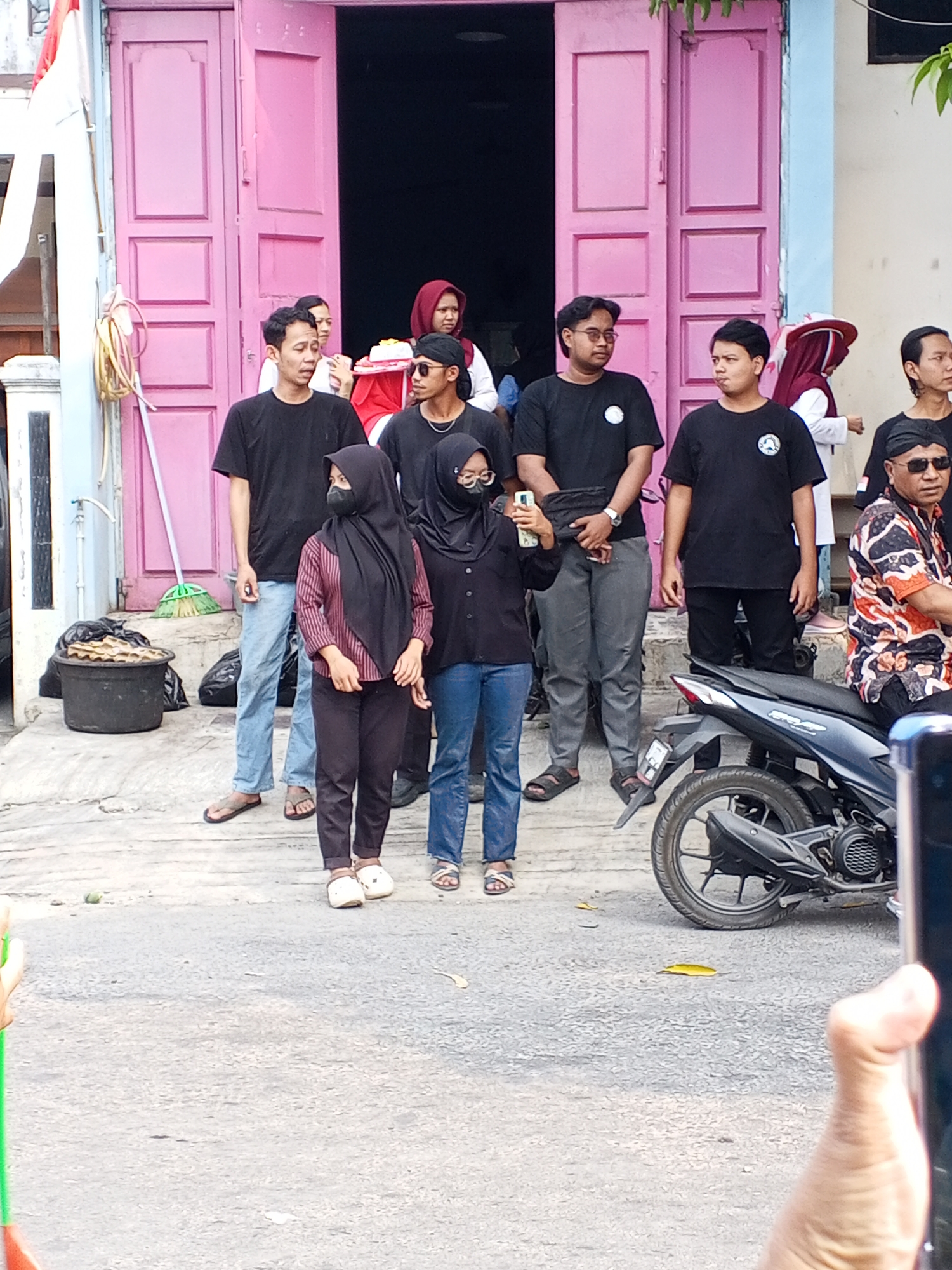 A group of people stand outside a building with bright pink double doors in the city of Ciruas, Indonesia. A flag with red and white stripes is attached to a pole on the left side of the building. The people are dressed casually, and most are wearing dark-colored t-shirts. Some are looking at the camera, while others are engaged in conversation or looking away. One person in the foreground is holding a smartphone. A motorcycle is parked to the right of the group. The ground is paved and appears to be a street or walkway. The overall scene suggests an informal gathering or event.
