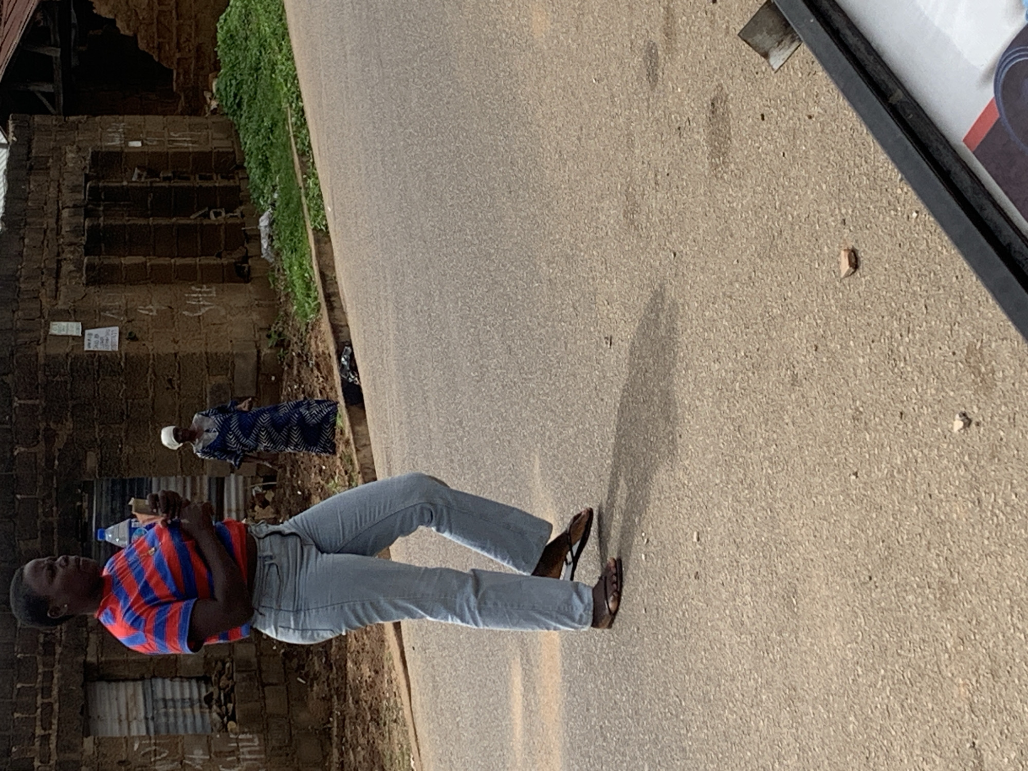In an outdoor scene on an asphalt road in Jos, Nigeria, a woman wearing a red and blue striped t-shirt and light blue jeans stands holding a bottle of water and a small object. Her body is angled away from the camera, and she is looking to her right. Behind her, another woman dressed in a blue and white patterned dress and a white headscarf stands near a dilapidated brick building with corrugated metal roofing. The building has several open window-like structures and appears to be in a state of disrepair. Green grass grows along the edge of the road, and there are various small stones and debris scattered on the asphalt. The lighting suggests it is daytime, with shadows cast by the people and structures.