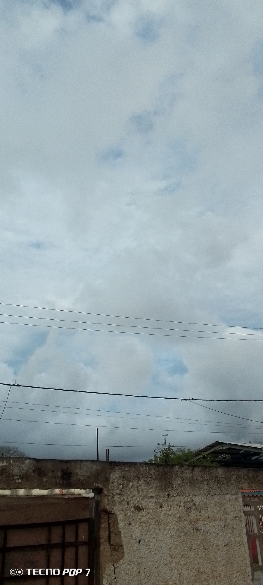 The image shows a gray, cloudy sky above a dilapidated wall in Altagracia, Venezuela. Multiple electrical wires are strung across the sky. The wall, made of textured concrete, appears aged and cracked, with a section missing on the left revealing a dark, metallic gate. Some green foliage is visible growing over the wall on the right side, along with a portion of a roof. The bottom left corner of the image displays the text "TECNO POP 7" in white lettering, indicating the device used to capture the photograph. There are no people visible in the scene.