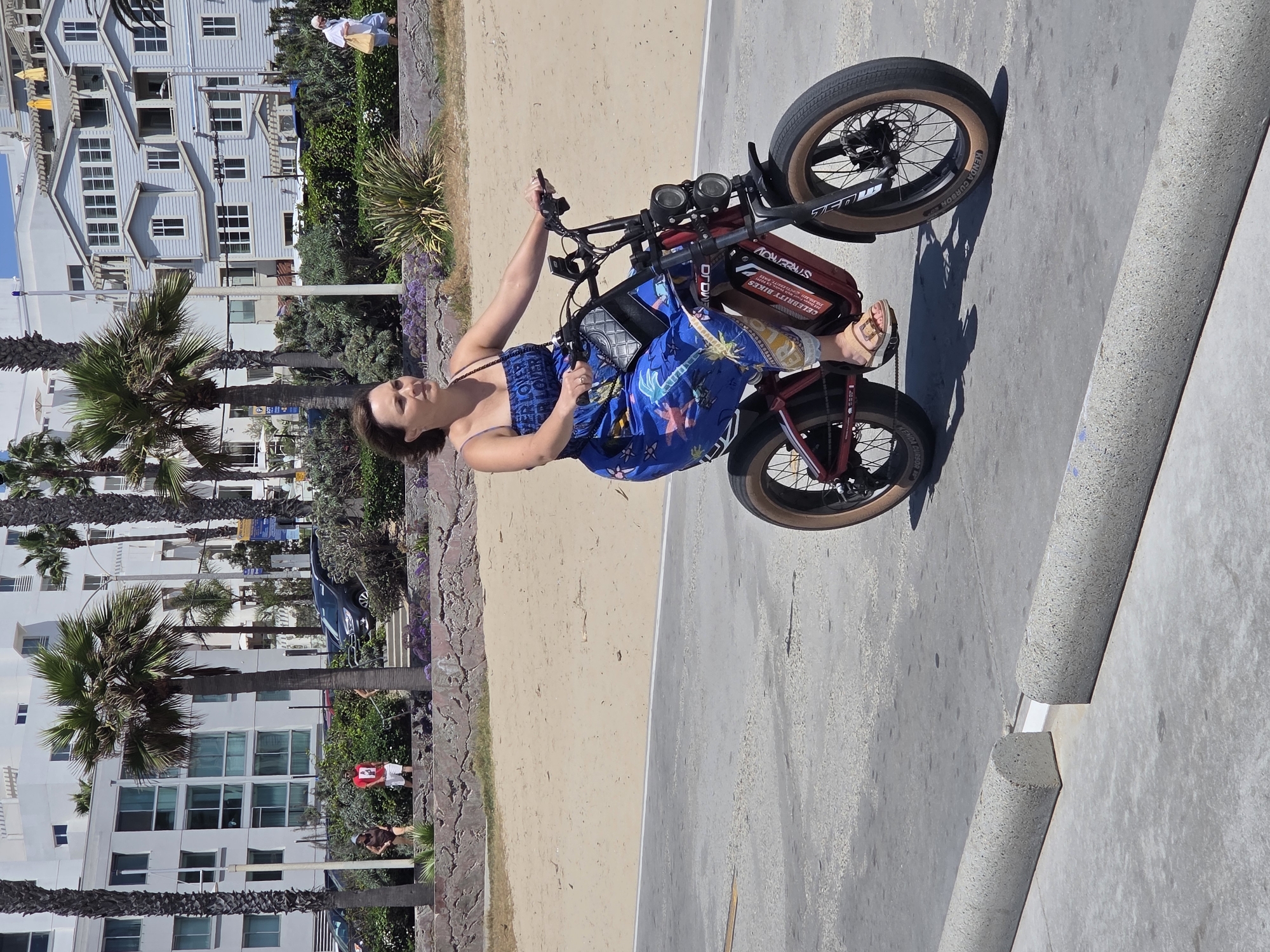 In Santa Monica, California, a woman is riding an electric bicycle on a paved path near a beach. The woman is wearing a blue spaghetti strap top and a long blue patterned skirt. She has brown hair and is wearing sandals. She is carrying a small quilted purse over her shoulder. The electric bicycle is red and black with thick tires and two headlights. The background shows a sandy beach with palm trees, a stone wall, and buildings. A dark SUV is parked on a street in the distance. The paved path has concrete curb stops.