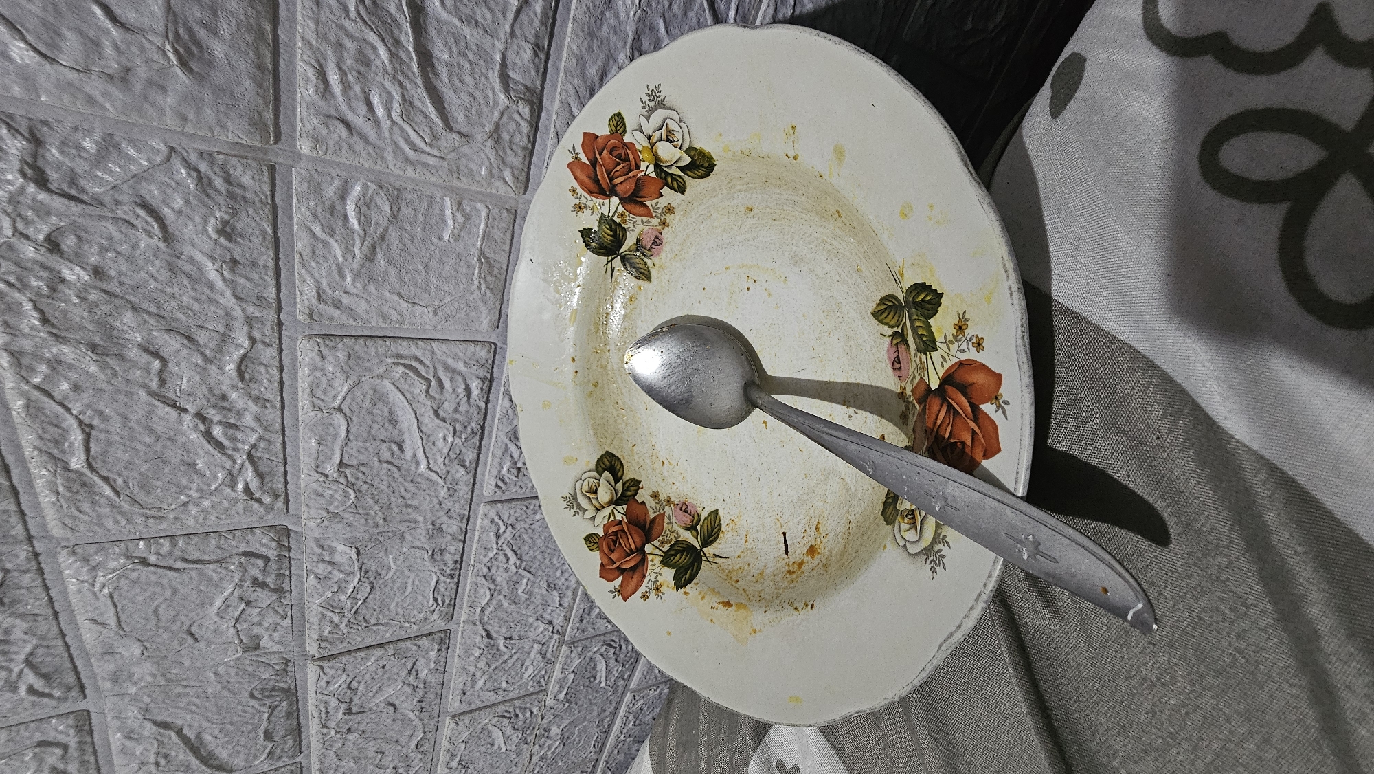 An overhead, slightly angled shot shows a used plate with a spoon resting on it. The plate is white with a floral pattern of red and white roses and green leaves. The plate has food residue on it and appears to have been used. The spoon is a standard metal spoon. The plate and spoon are positioned on a surface covered with a striped cloth. In the background, a textured grey brick wall is visible. The scene appears to be indoors, likely on a table. The lighting suggests it is likely evening or a dimly lit room.