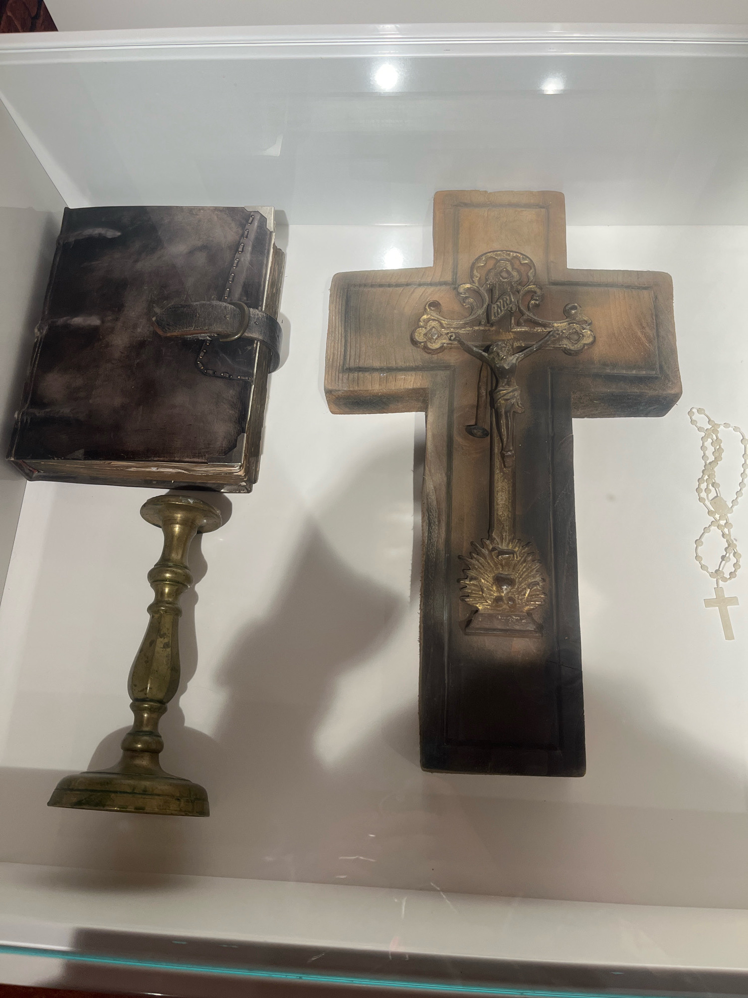 The image displays religious artifacts within a glass display case. On the left, a dark, leather-bound book rests on a brass candlestick. The book appears old, with a strap closure and metal corner protectors. To the right, a large wooden cross with a metal crucifix depicting Jesus is prominently featured. To the right of the cross, a string of white rosary beads hangs, with a small cross at the end. The background is white, suggesting a museum or exhibit setting. This scene is located in the Chapel of the Three Saints at the Lychakiv Cemetery in Lviv, Ukraine.