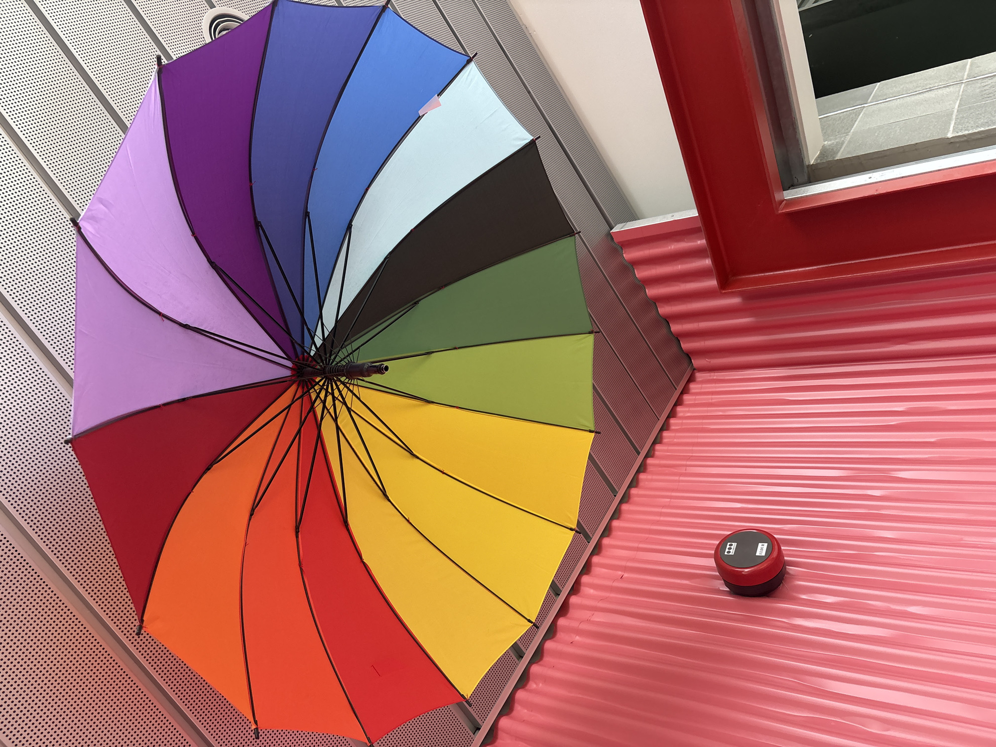 The image shows a colorful rainbow umbrella suspended from a perforated ceiling. Below the umbrella, a bright pink corrugated wall is visible, with a red fire alarm mounted on it. To the right, a red door frame opens to a glimpse of a tiled interior. The scene appears to be indoors, possibly a public space or a creatively decorated area.