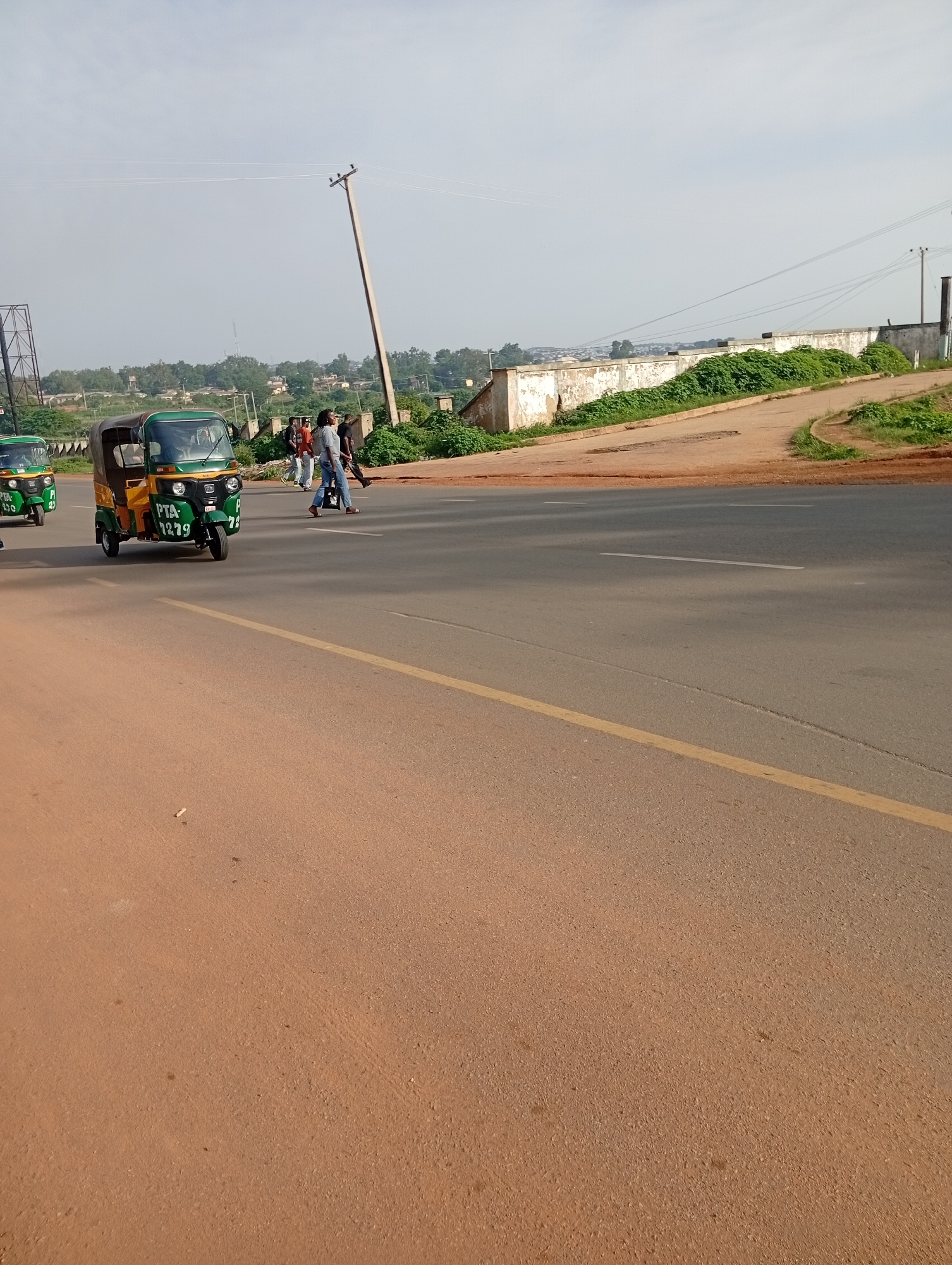 The image depicts a street scene in Rafin Par, Nigeria. In the foreground, a paved road with a yellow dividing line is visible, with a reddish-brown shoulder on the left. In the midground, a green and yellow auto-rickshaw, identified by the license plate "PTA 1279", is traveling on the road. Another auto-rickshaw, partially visible, is to the left of the first. Several pedestrians are crossing the road in the distance. Beyond the road, there is a concrete wall overgrown with green vegetation, and further in the background, a cityscape with buildings and trees is visible under a hazy sky. A utility pole stands prominently near the center of the image.