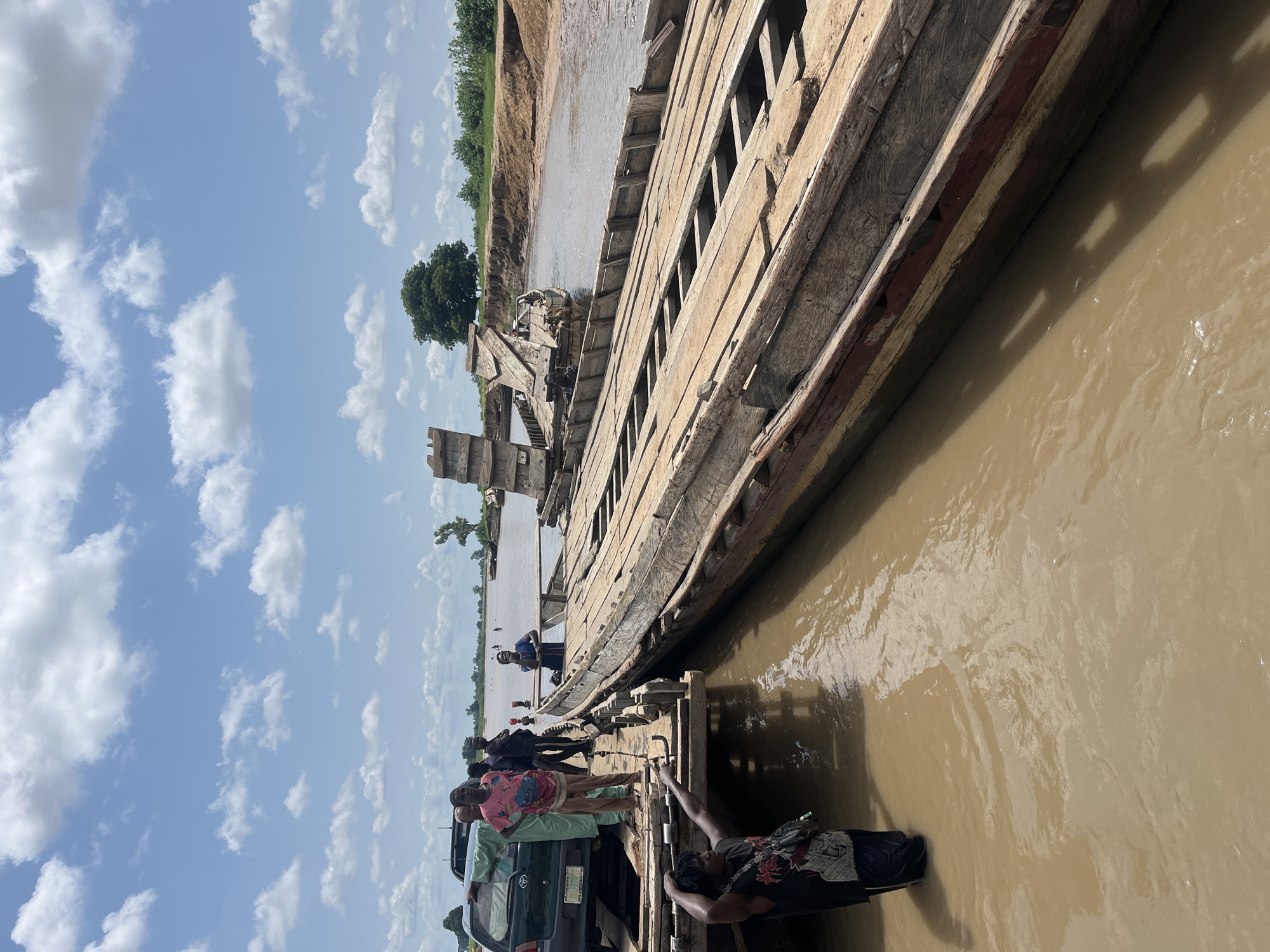 A group of people are gathered around a damaged wooden ferry on a muddy river. A green SUV is parked on the ferry's deck, suggesting an attempt to transport it across the river. The ferry appears to be made of multiple connected wooden boats, some of which are significantly broken and dilapidated. The riverbank in the background is lined with vegetation, and trees are visible further inland. The sky is blue with scattered white clouds. The scene is set in Nemnei, Nigeria.