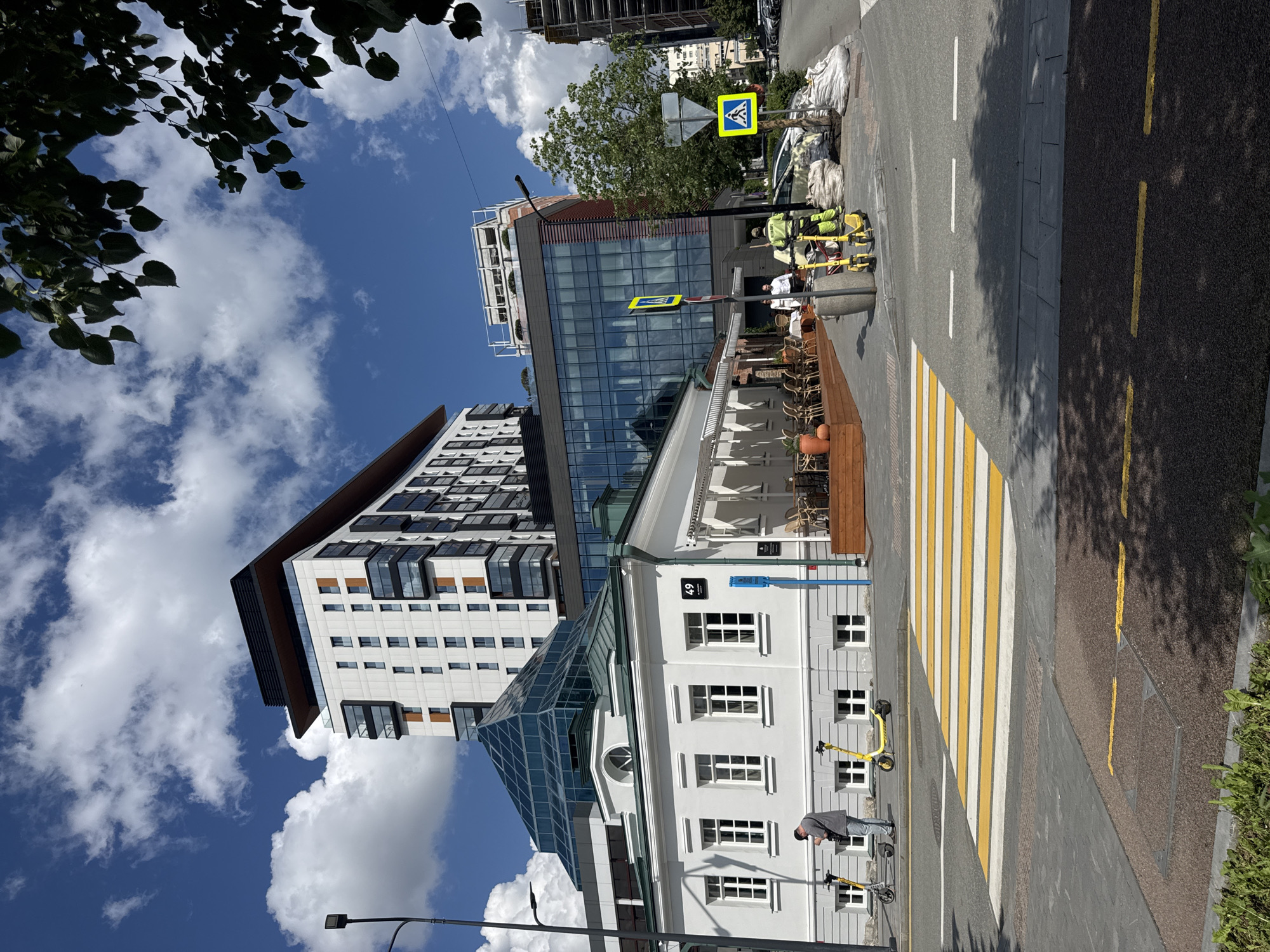 The image captures a street scene in Yekaterinburg, Russia, likely near the intersection of 49, Malysheva Street and Vaynera Street, given the building number visible. In the foreground, a pedestrian crossing with yellow and white stripes is prominent on the asphalt road. Two yellow electric scooters are parked on the sidewalk to the left, near a low-rise white building with multiple windows and a light gray base. A man in a gray t-shirt and blue jeans stands next to one of the scooters, looking down at his phone. Behind this building, a modern multi-story building with a glass pyramid-shaped roof extension is visible. Further back, a taller, modern apartment building with white, beige, and black facade elements rises into the bright blue sky filled with fluffy white clouds. To the right, another modern building with a glass facade is present. On the sidewalk near the glass-fronted building, two individuals, one in a bright yellow uniform and the other in white, are visible. The person in yellow is working with equipment, including yellow electric scooters and some large white bags, possibly for landscaping or construction. Road signs, including a pedestrian crossing sign with a blue background and a white silhouette of a person, are also present. The environment suggests an urban setting with a mix of historical and contemporary architecture.