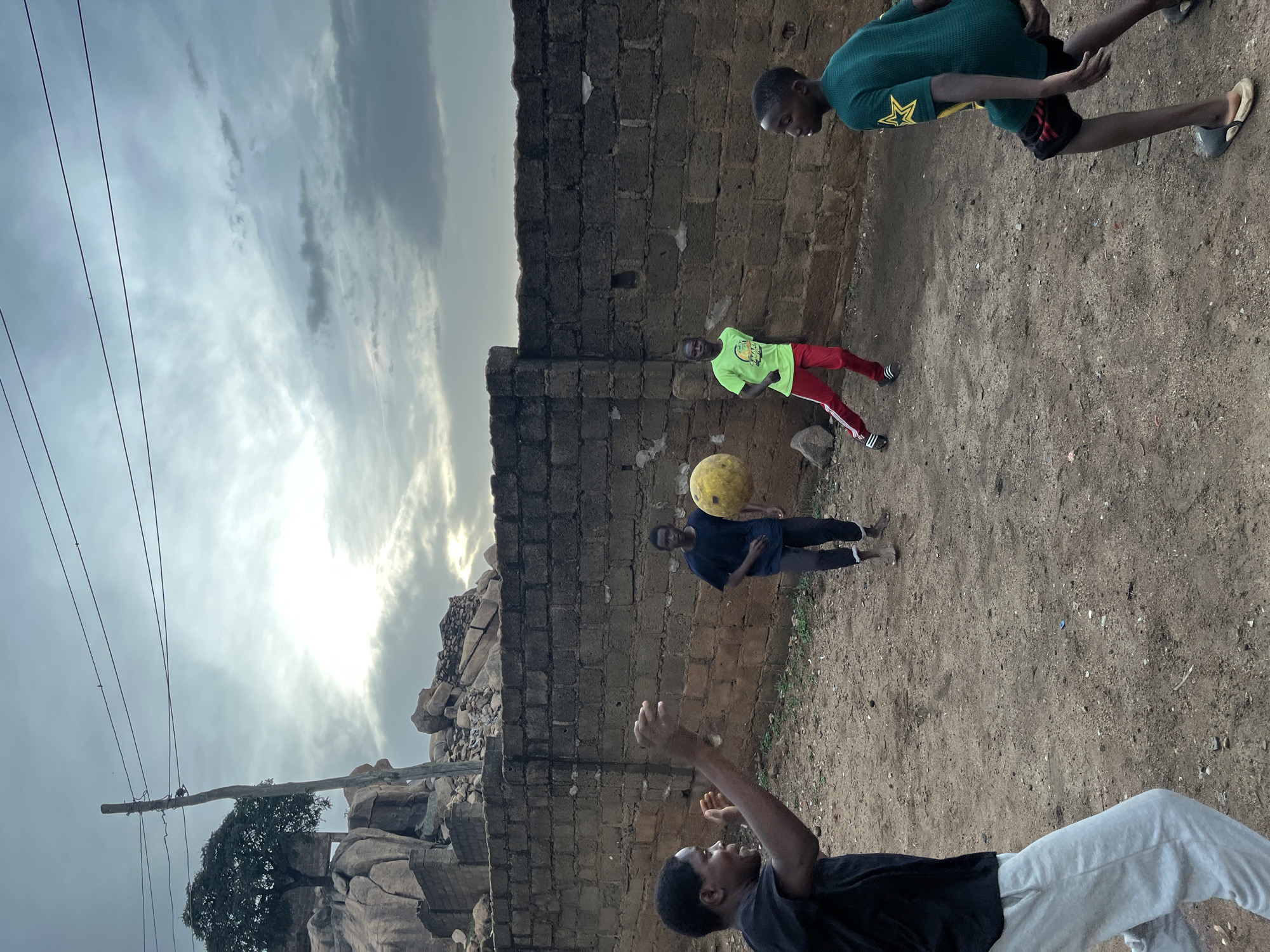 Five young men are outside near a brick wall. One man near the bottom of the image is wearing a black t-shirt and white pants and has his arms up. The next man is wearing a dark blue t-shirt and dark pants, and there is a yellow ball in the air near him. The next man up is wearing a bright green t-shirt and red pants. The next man up is wearing a green t-shirt with a gold star. The background shows a cloudy sky and a rock formation. It is outdoors in Jos, Nigeria.