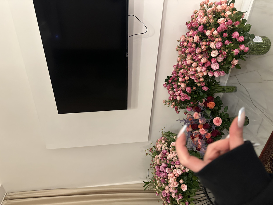 The scene depicts an interior room, likely a modern living space or lobby within Yerevan, Armenia. The specific location within the city is not identifiable. In the background, a large, black Grundig television is wall-mounted on a white panel, itself affixed to a light-colored wall. To the left, a beige curtain hangs. The foreground is dominated by multiple large, dense bouquets of fresh cut flowers, predominantly small pink and peach spray roses, with some arrangements also containing darker red and purple blooms. These bouquets are displayed in various clear glass vases on a low, reflective, light-colored surface, possibly marble. A human left hand, adorned with long, light-colored manicured nails and wearing a dark long-sleeved garment, is visible in the lower-left corner, positioned in a pinching gesture with the thumb and index finger. A portion of a dark, patterned rug is visible beneath the hand. No other individuals or actions are depicted.