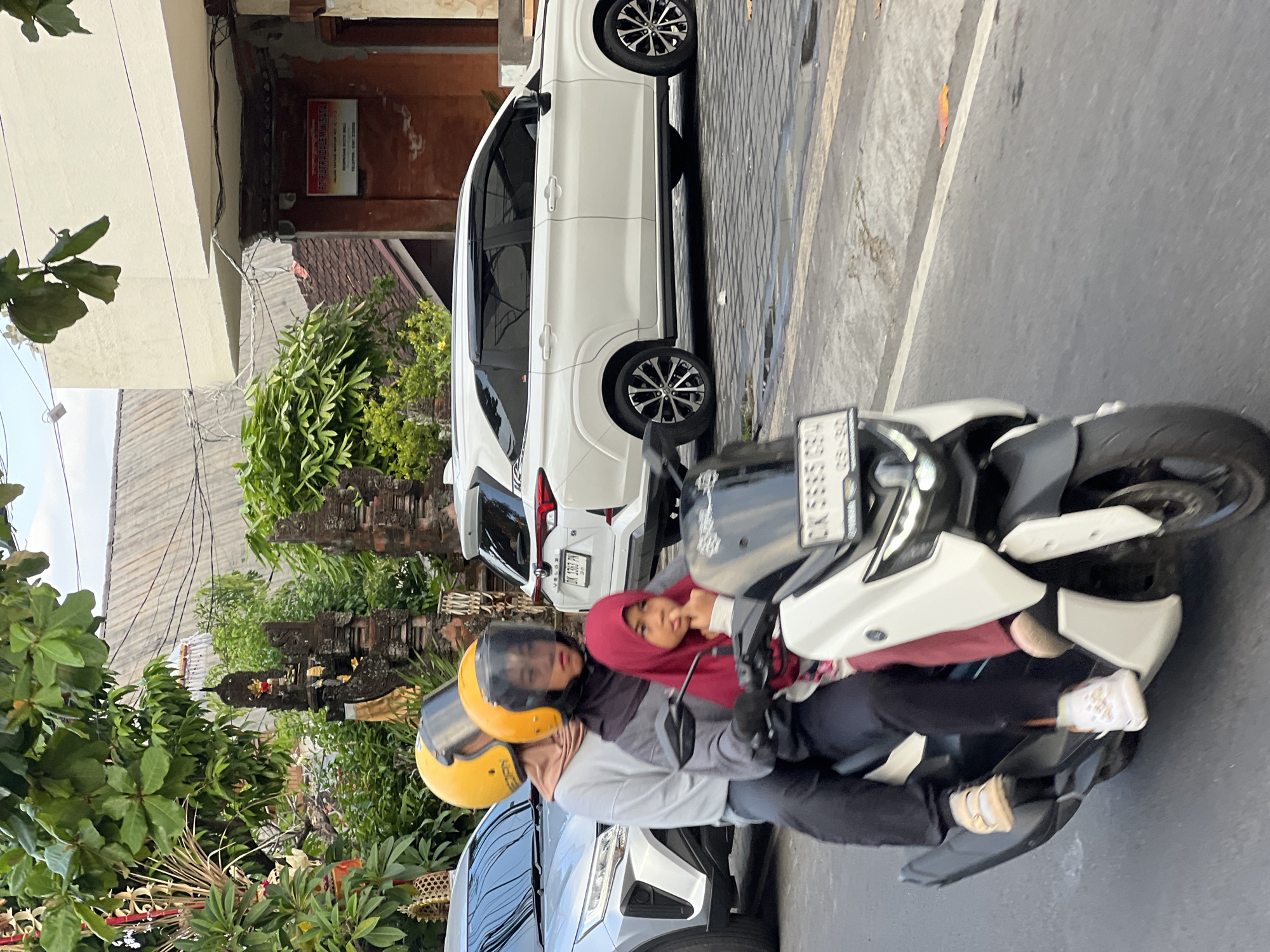 This is a medium shot photograph of three people riding on a white and black scooter in Denpasar, Indonesia. The scooter is moving on a paved road. The person in the front is wearing a dark grey long-sleeved shirt, black pants, a black headscarf, and a yellow helmet. They are operating the scooter. The person in the middle is wearing a light grey long-sleeved shirt, black pants, and a yellow helmet. The person in the back is wearing a red hijab and a light-colored top. They are holding onto the person in the middle. The scooter has a license plate that reads "DK 5665 GBW". In the background, there is a white SUV, a silver car, and traditional Balinese temples. Lush green trees and buildings are also visible. The sky is partly cloudy. The lighting suggests it is daytime, likely late afternoon.