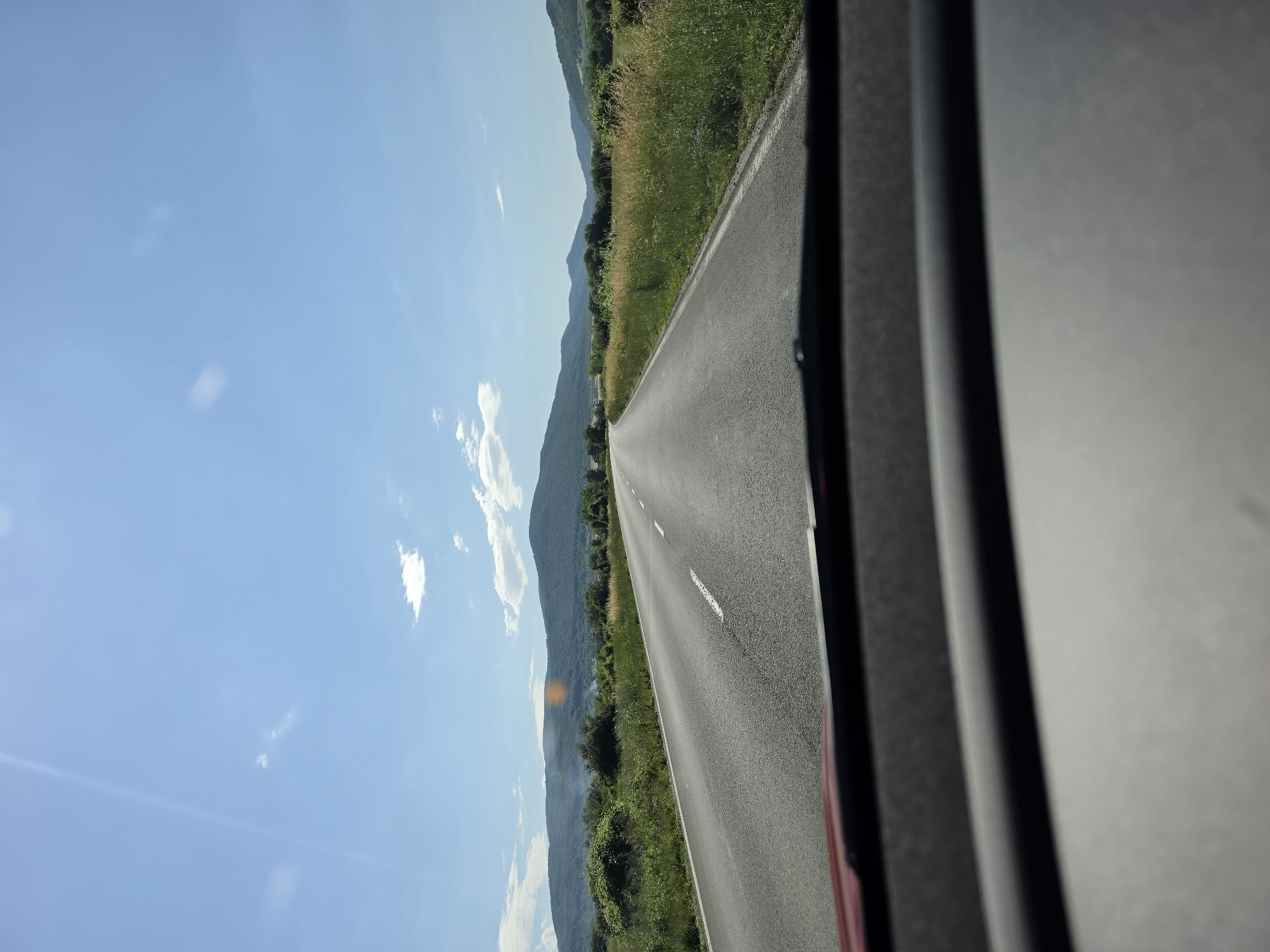 The image depicts a rural or semi-rural landscape observed from the interior of a vehicle, looking forward through the windshield. A two-lane asphalt road, dry and dark gray, extends from the immediate foreground into the distant background, featuring a broken white center line and a solid white edge line on the right. The road appears to gently curve to the right in the distance.
On both sides of the road, there are shoulders leading to dense green and yellowish-green vegetation, primarily tall grasses and bushes, indicating a natural or uncultivated environment. Beyond the immediate roadside foliage, rolling hills and mountains covered in lush green deciduous trees and shrubs dominate the mid-ground and background, extending across the horizon. A faint plume of light smoke or haze is visible above the vegetation on the left side, near the base of the leftmost mountain range.
The sky is clear and bright blue with scattered, wispy white cumulus clouds. Intense sunlight from an elevated position causes visible lens flares or glare in the upper left and central right portions of the sky, indicating a sunny day. The lower portion of the image displays the vehicle's dashboard, dark gray and black, slightly out of focus. No individuals or other vehicles are visible on the road or in the surrounding landscape. The scene is located in Riahovtsite, Bulgaria.