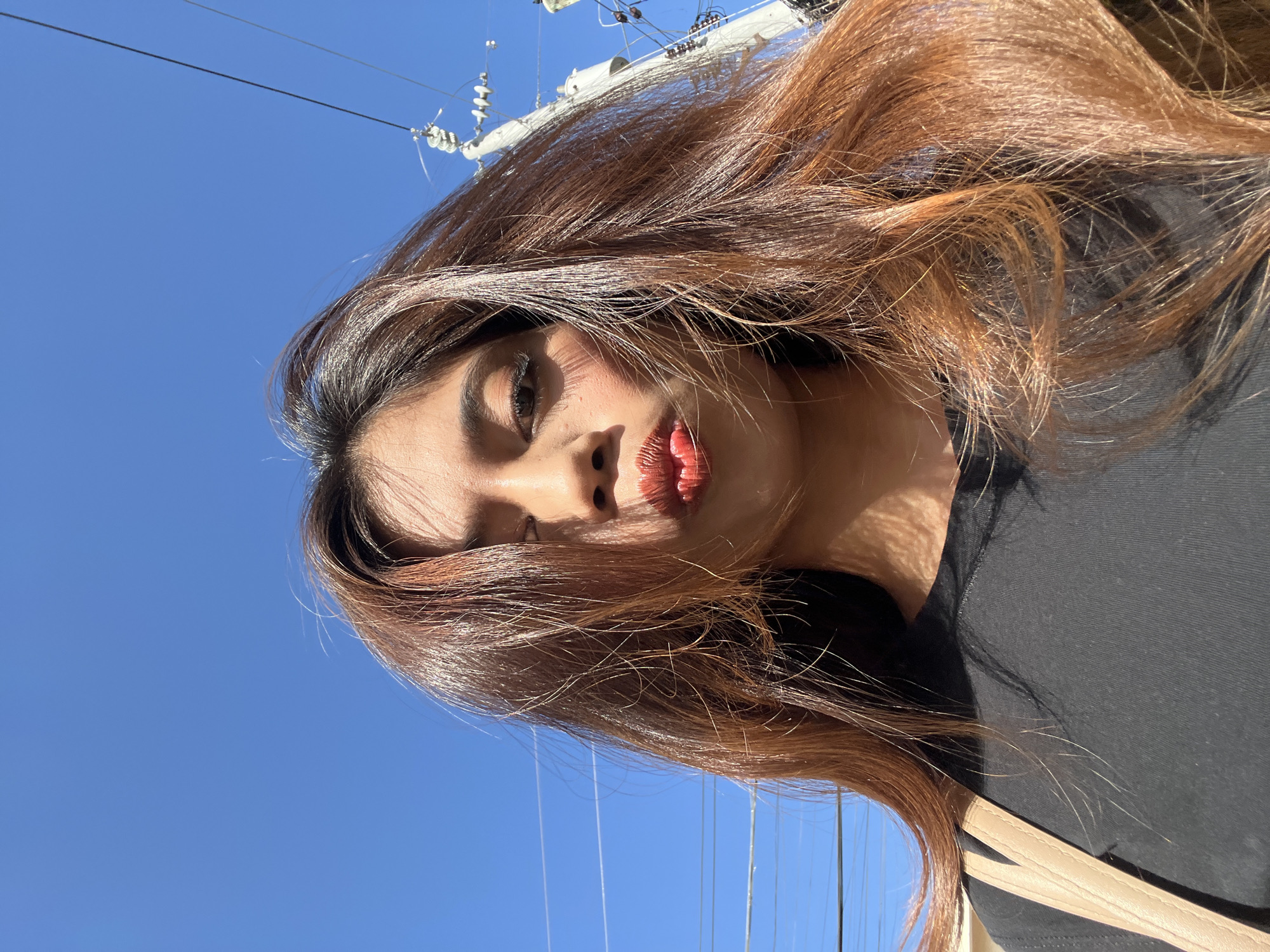 A close-up, low-angle selfie features a young woman with long, brown hair blowing in the wind. She is wearing a black t-shirt and a light-colored strap from a bag is visible on her left shoulder. Her lips are puckered and highlighted with bright red lipstick. The background is a clear blue sky with a few utility poles and power lines visible, suggesting an outdoor urban setting. The lighting indicates it is daytime, with sunlight casting shadows on her face and hair. The scene is likely situated in an open area within Davao City, Philippines, given the presence of utility infrastructure typical of the region.