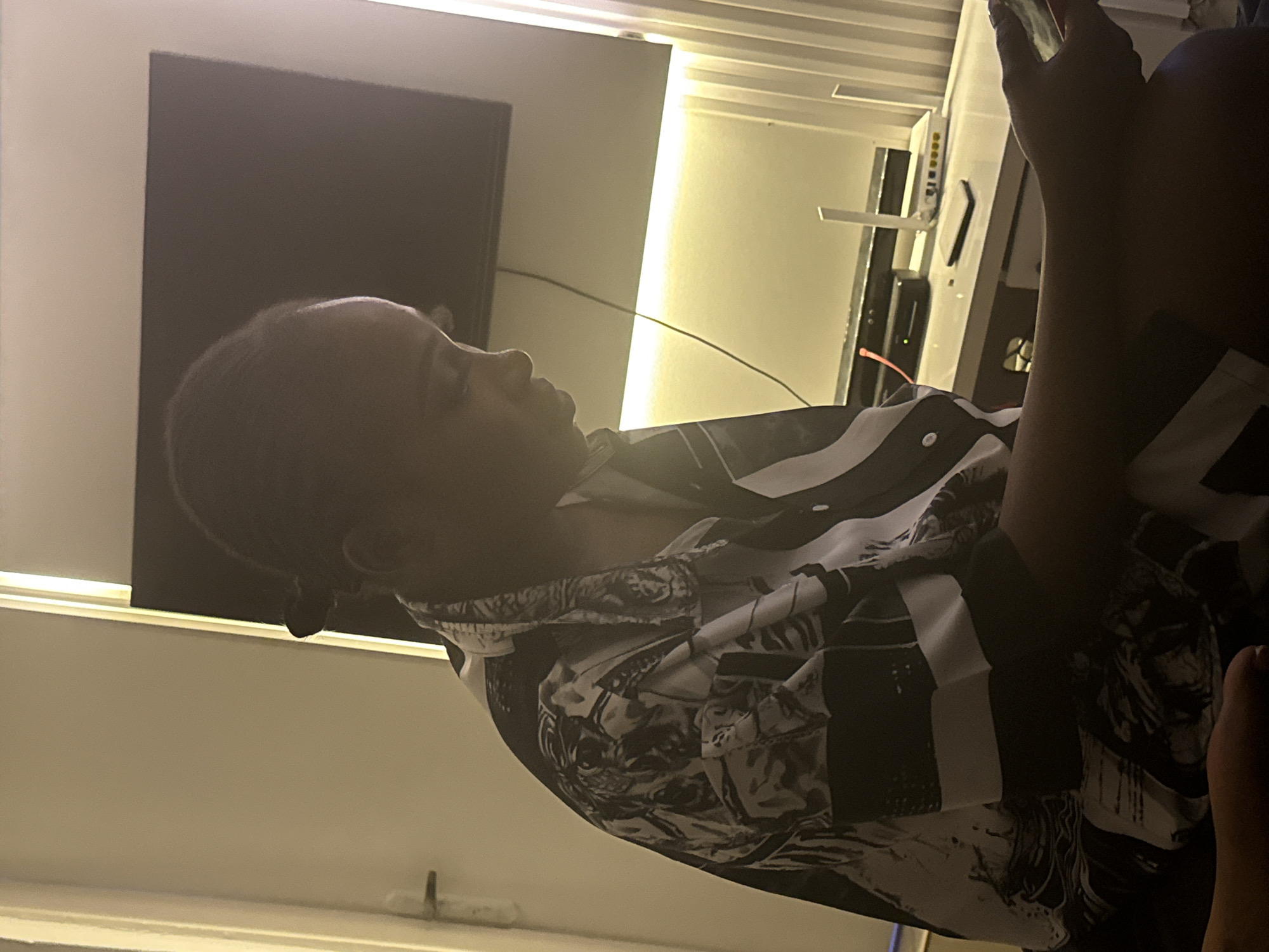 The image depicts a side profile of a young Black woman seated indoors, engrossed in her phone. She is wearing a black and white patterned button-up shirt. Her hair is pulled back in a braid. In the background, a television screen is visible, along with a desk containing a router and other electronic devices. The room is dimly lit, with artificial lighting highlighting the wall and the woman's face and clothing. The overall impression is that of a casual, indoor setting, possibly a living room or bedroom. The location within Abuja, Nigeria, cannot be determined from the visual information alone.