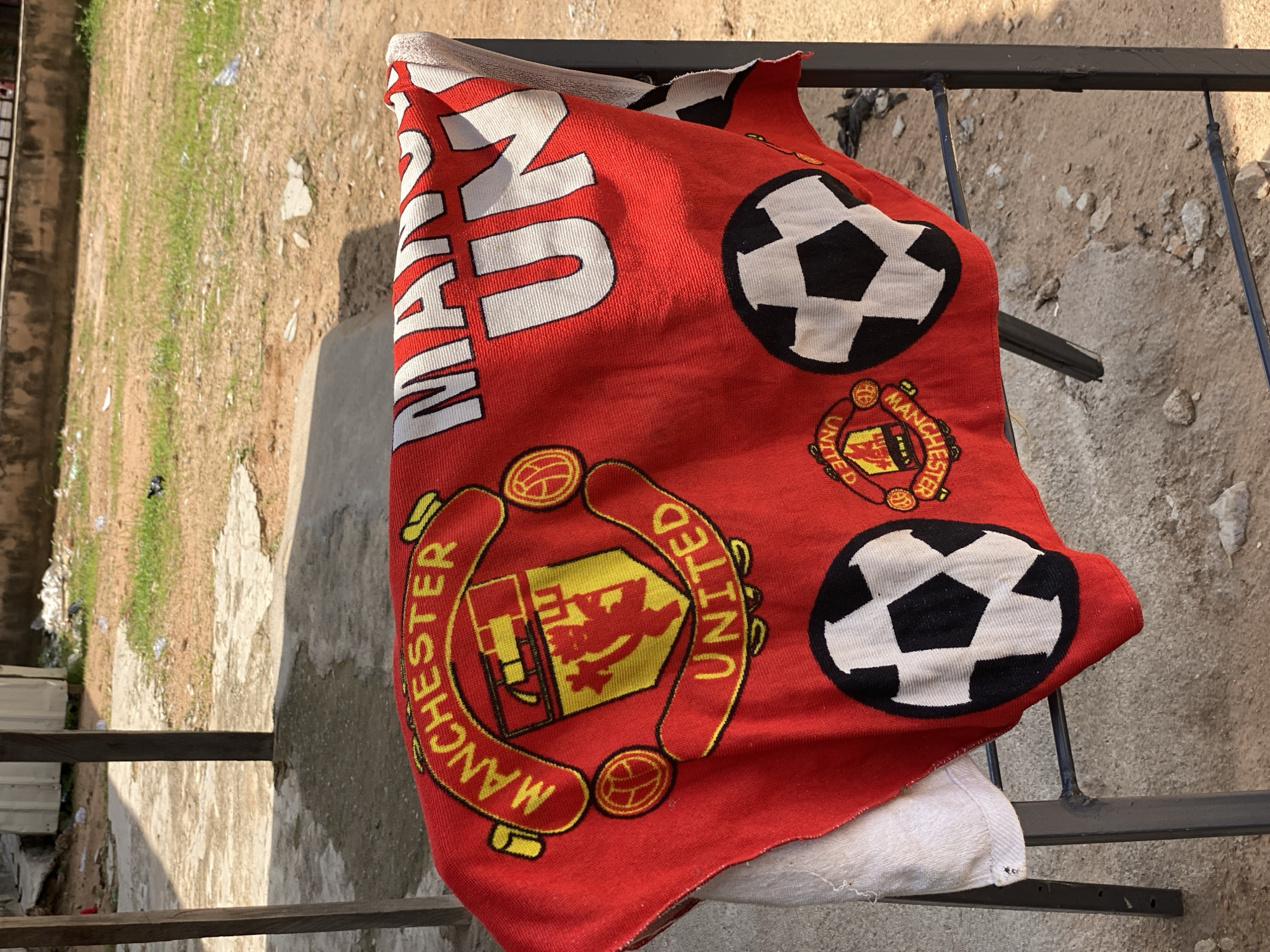 A red Manchester United football club towel is draped over a black metal rack in an outdoor setting. The towel features the club's crest and a soccer ball graphic. The background shows a rough, unpaved ground with scattered rocks and dried vegetation, indicating a rural or semi-developed area. A concrete structure and wooden posts are visible in the background, along with some discarded plastic bottles and debris, suggesting an unkempt environment. The overall impression is one of a dry, dusty outdoor space, possibly on the outskirts of a town like Jalingo, Nigeria. The image is taken during daylight with natural lighting.