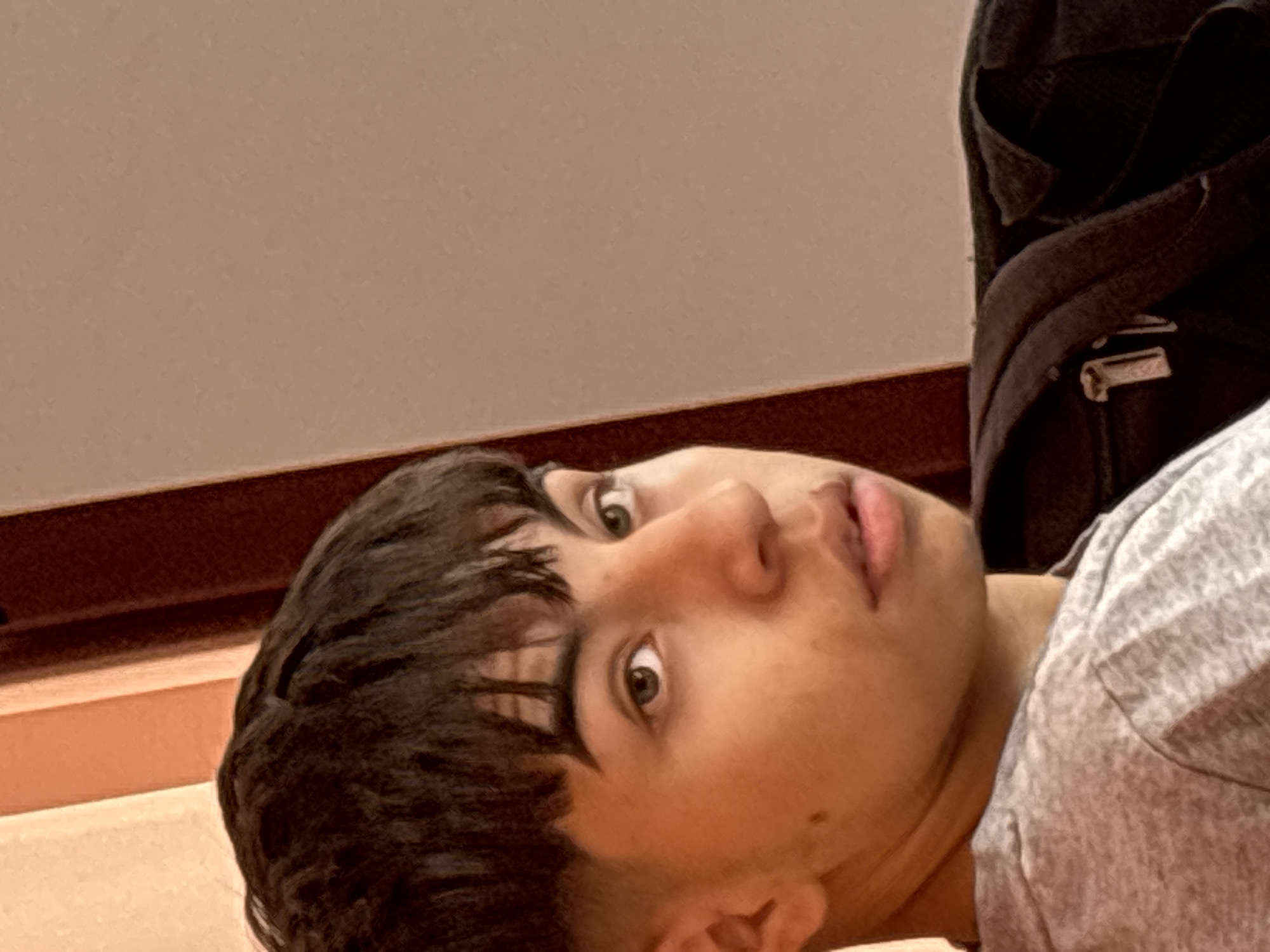The image presents a close-up, angled shot of a young person, likely male, with their head tilted sharply to the right. The individual has dark, textured hair that falls across their forehead, and light-colored eyes, possibly green or hazel, that are wide open and looking directly at the viewer. Their mouth is slightly agape, contributing to an expression that appears curious or perhaps a little surprised. A small mole is visible on their right cheek.
They are wearing a light gray t-shirt with a textured fabric. In the upper right corner of the frame, a dark-colored backpack or bag is visible, suggesting an indoor setting, possibly a school or a casual public environment. The background consists of a plain, light brown or tan wall, with a darker reddish-brown horizontal band or trim running along the lower portion of the wall, behind the person's head.
No specific activity is explicitly taking place other than the act of being photographed. The unusual camera angle gives the impression of a casual, possibly candid or selfie-style shot. There is no visible text or specific location cues beyond an indoor setting. The time of day and weather are not discernible.