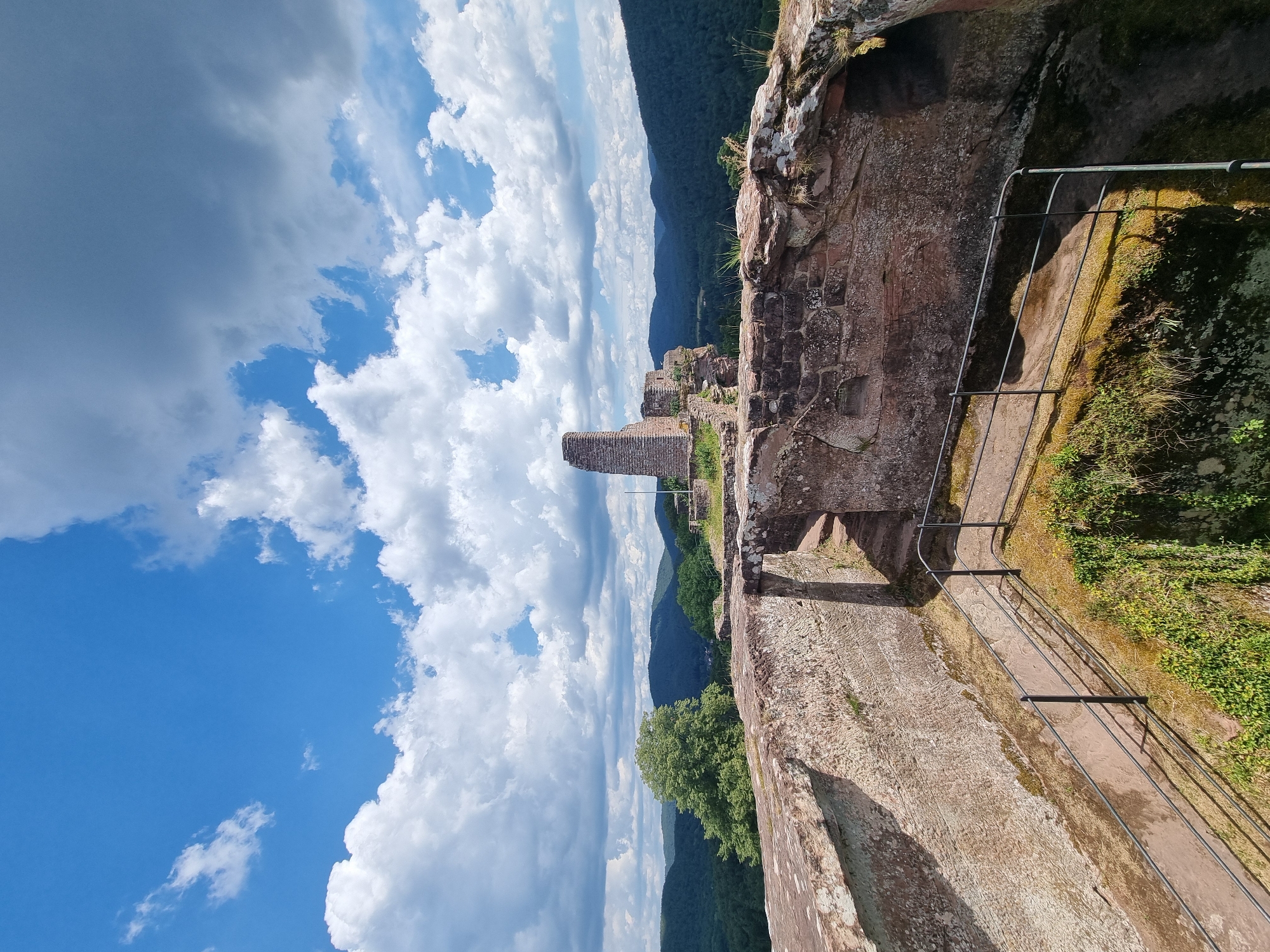The image shows the ruins of a castle on a hilltop overlooking a forested valley under a bright blue sky with scattered white clouds. The foreground features the stone walls of the castle, weathered and overgrown with moss and grass. A metal railing guides visitors along a path next to a steep drop. In the background, the remnants of a tower and other stone structures stand against the backdrop of rolling green hills. The scene is set in the morning or afternoon, as indicated by the strong sunlight casting shadows. This is the Dahner Schlossberg, a historical landmark in Dahn, Germany, known for its ancient fortifications and scenic views. No people are visible in the image.
