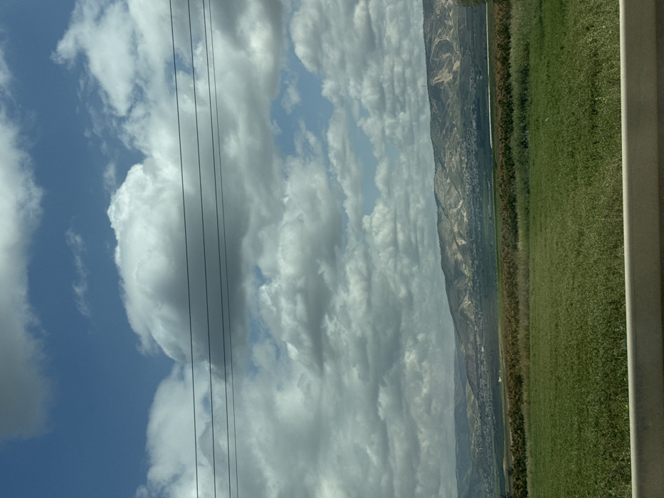 The image presents a wide-angle landscape view captured from a vehicle, looking across an agricultural plain towards distant hills under a partly cloudy sky within the Emek HaMaayanot Regional Council, Israel. The upper half of the frame shows a blue sky dominated by numerous white and grey cumulus clouds, with clear blue patches visible between them. Four thin, dark power lines stretch horizontally across the upper-middle section of the image. The immediate foreground is occupied by a light-colored, horizontal barrier, likely a vehicle's dashboard or a road guardrail. Beyond this, a vast expanse of vibrant green cultivated fields fills the lower-mid ground. Farther back, a band of denser, darker vegetation, possibly mature crops or natural flora, precedes a valley floor where small clusters of buildings, indicative of settlements, are discernible. The background features a continuous range of light-colored hills or mountains, displaying varying patterns of vegetation and exposed earth. No individuals or specific activities are observed.