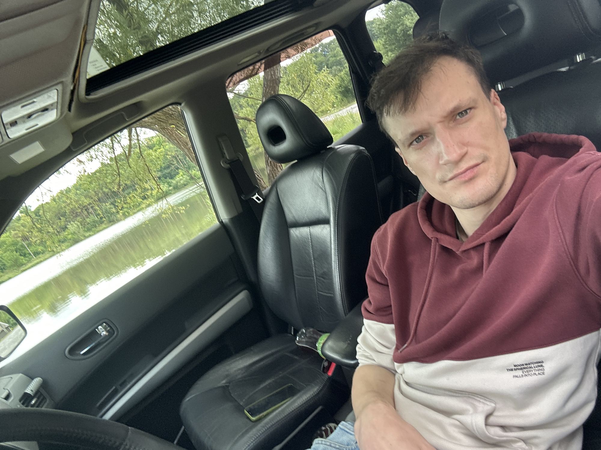 The image depicts a person reclining in the passenger seat of a car. The person is wearing a maroon and cream colored hoodie. The car's interior is visible, including black leather seats and a sunroof. Outside the car, a body of water and trees are visible, suggesting a natural, possibly rural setting. The provided crops focus on details of the car's interior, the person's clothing, and their facial features. There is no indication that the image was taken in the city of Makiv, Ukraine.