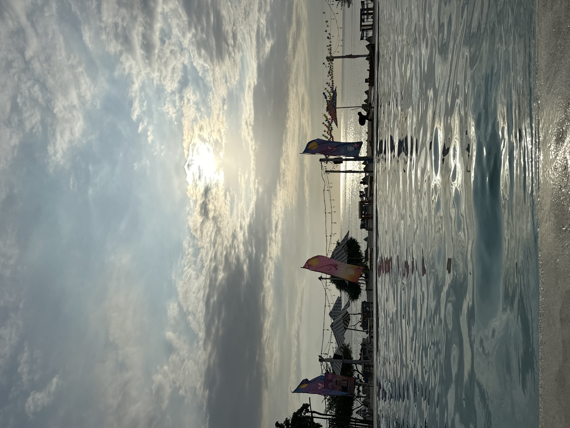 The photograph captures a waterfront scene in an unidentified location in Indonesia. The foreground features the rippled surface of water, possibly a pool or bay. Along the edge of the water are several colorful flags and beach umbrellas, suggesting a recreational area. In the distance, the sun glints on the water, creating a bright reflection. The sky is filled with numerous clouds, and the sun is partially obscured, casting a soft light over the scene. Several figures can be seen in the background.