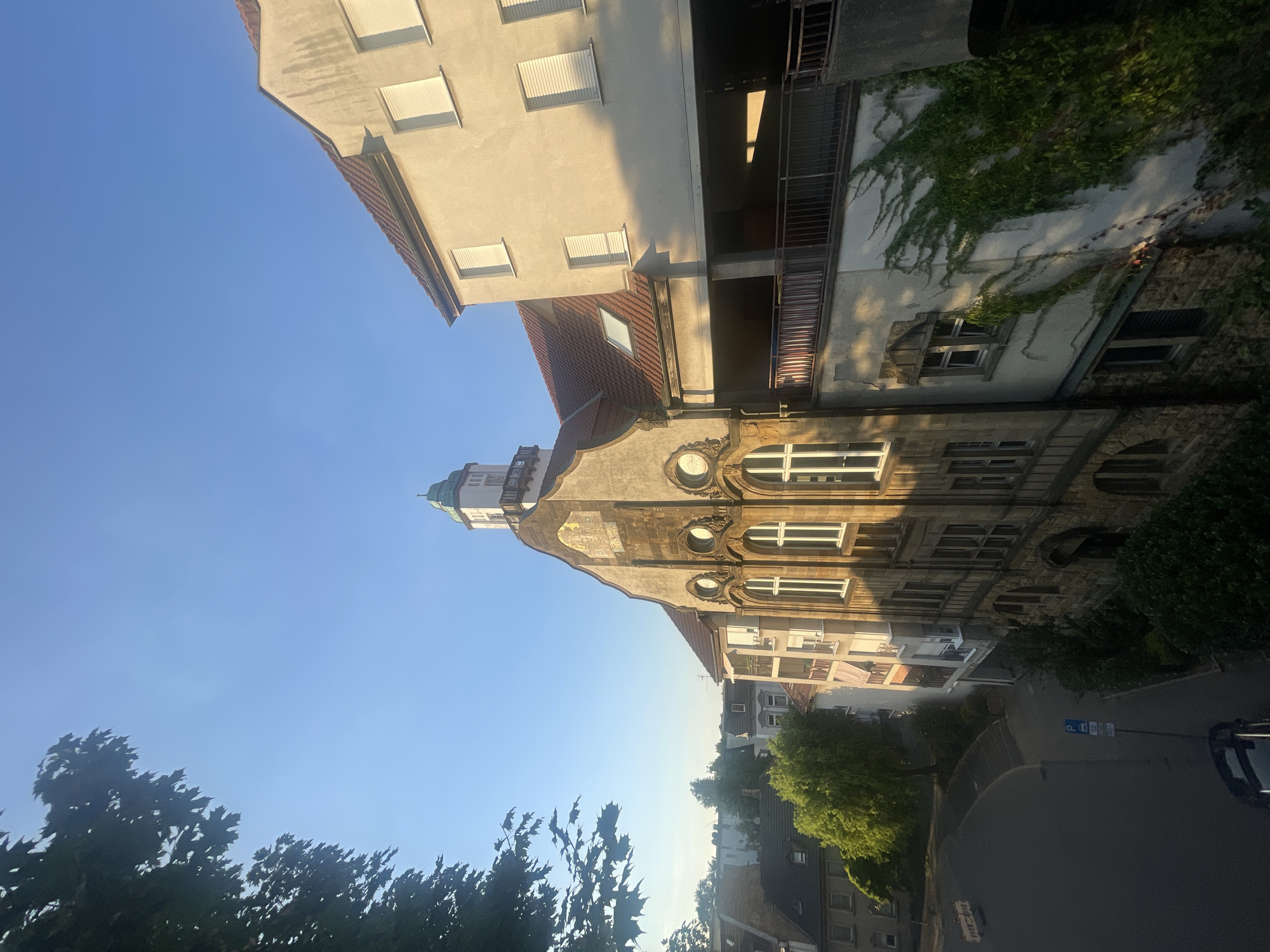 The image shows a view of buildings in Iserlohn, Germany, under a clear blue sky. In the foreground, a tree with dark green leaves is visible on the left. Dominating the center of the frame is an old, multi-story building with a distinctive facade featuring stone ornamentation and arched windows. This building has a pitched roof and a small tower with a green, pointed top. To the right of this building is a lighter-colored, more modern structure with multiple windows covered by white shutters. A balcony or walkway with a railing separates the two buildings at a mid-level. Ivy is growing up the side of the lighter building. Below these buildings, a street curves to the left, with a parked car and a blue parking sign visible. Further in the background, other buildings and trees can be seen, suggesting a residential or mixed-use urban area. The lighting indicates it is likely morning or late afternoon due to the strong shadows cast.