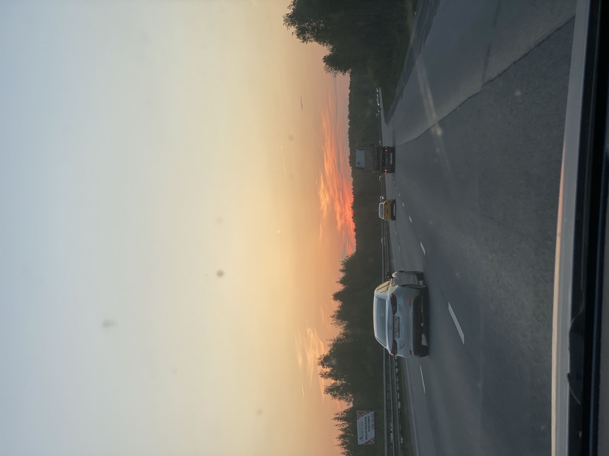 The image depicts a highway scene during a sunset, likely taken from inside a vehicle. A grey car is visible in the right lane, with its license plate partially visible as "P782CM96." Further ahead on the highway, there is a yellow car, and a truck. The sky is a gradient of orange and yellow, indicating sunset. The trees line the highway on both sides. A road sign is barely visible at the bottom left corner of the frame, and the highway is divided by white lane markings. The overall impression is a scene of a typical highway journey at dusk. It is impossible to determine the city with the information given.