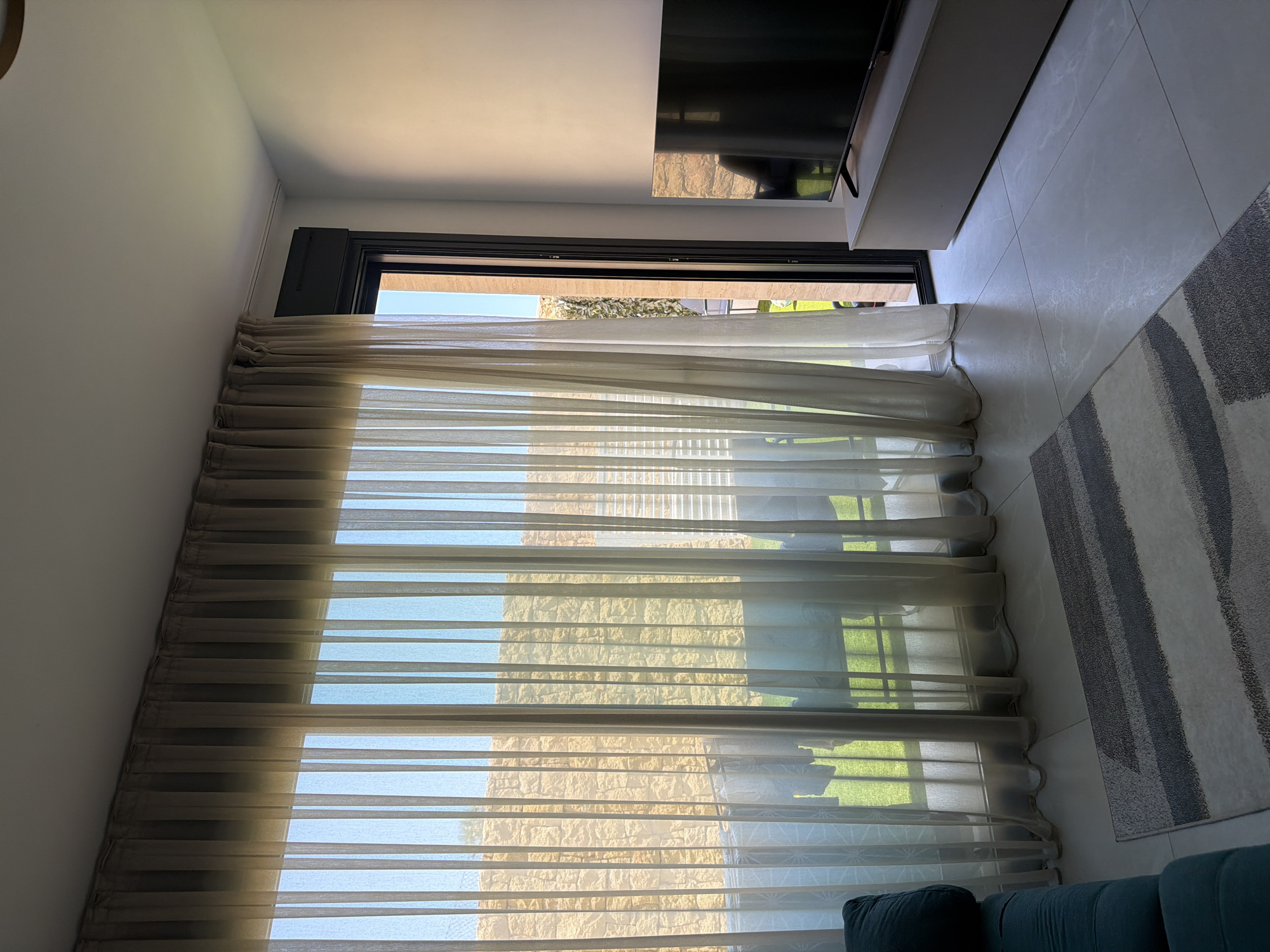 The image depicts a well-lit interior scene, likely a living room, in Finestrat, Spain. A large window with sheer, beige curtains dominates the left side of the frame, allowing sunlight to stream in and illuminate the room. Through the curtains, a stone wall and a green outdoor space, possibly a balcony or garden, are visible. To the right, a modern black television is placed on a light-colored entertainment unit. The floor is tiled with a light gray marble pattern. In the foreground, a grey and white striped rug is laid out. A portion of a teal sofa is visible in the lower left corner. The overall ambiance is bright and airy, suggesting a daytime setting. There are no people present in the scene.
