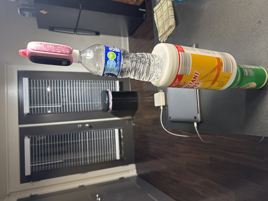 The scene is an interior view of a residence in Gainesville, United States of America. On a dark gray countertop in the foreground, a stack of four items is precariously balanced. From bottom to top, these items are: a green and white cylindrical container, a white "DISINFECTANT WIPES" container with a red and yellow label, a partially-filled clear plastic water bottle, and a pink and black electronic device resembling a vape, balanced upright on the bottle's cap. To the left of the stacked items, a closed silver laptop is connected via a white cable to a white power adapter. A folded plaid kitchen towel in green, white, and brown is on the countertop to the right. The background features dark hardwood flooring, two dark-colored doors with closed horizontal blinds, and a black trash can containing a pink liner. A portion of a black refrigerator is visible on the far right. No individuals are present in the image.