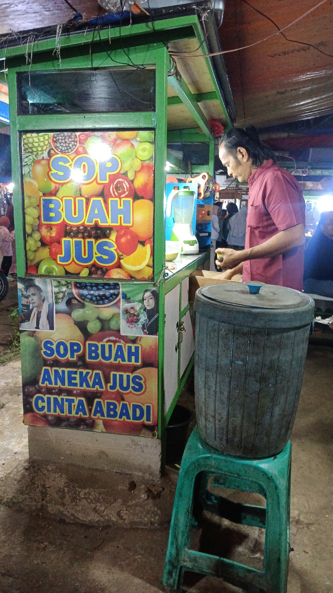 A man with dark, tied-back hair stands behind a food stall, wearing a maroon collared shirt. He is facing to the right, with his right hand holding a small blue object. In front of him, to the left, is a green and white food stall with signs advertising "SOP BUAH" and "BUAH JUS." The stall features colorful images of fruits. To the right of the stall, a large, gray, cylindrical container sits on a green plastic stool. The ground is a rough, concrete surface. In the background, other people are visible, some blurred. The scene appears to be outdoors, possibly a market, with overhead structures and wires suggesting a covered area. The overall lighting is dim, with some areas illuminated by artificial light. The text on the stall is in Indonesian. The location is Tajurhalang, Indonesia.