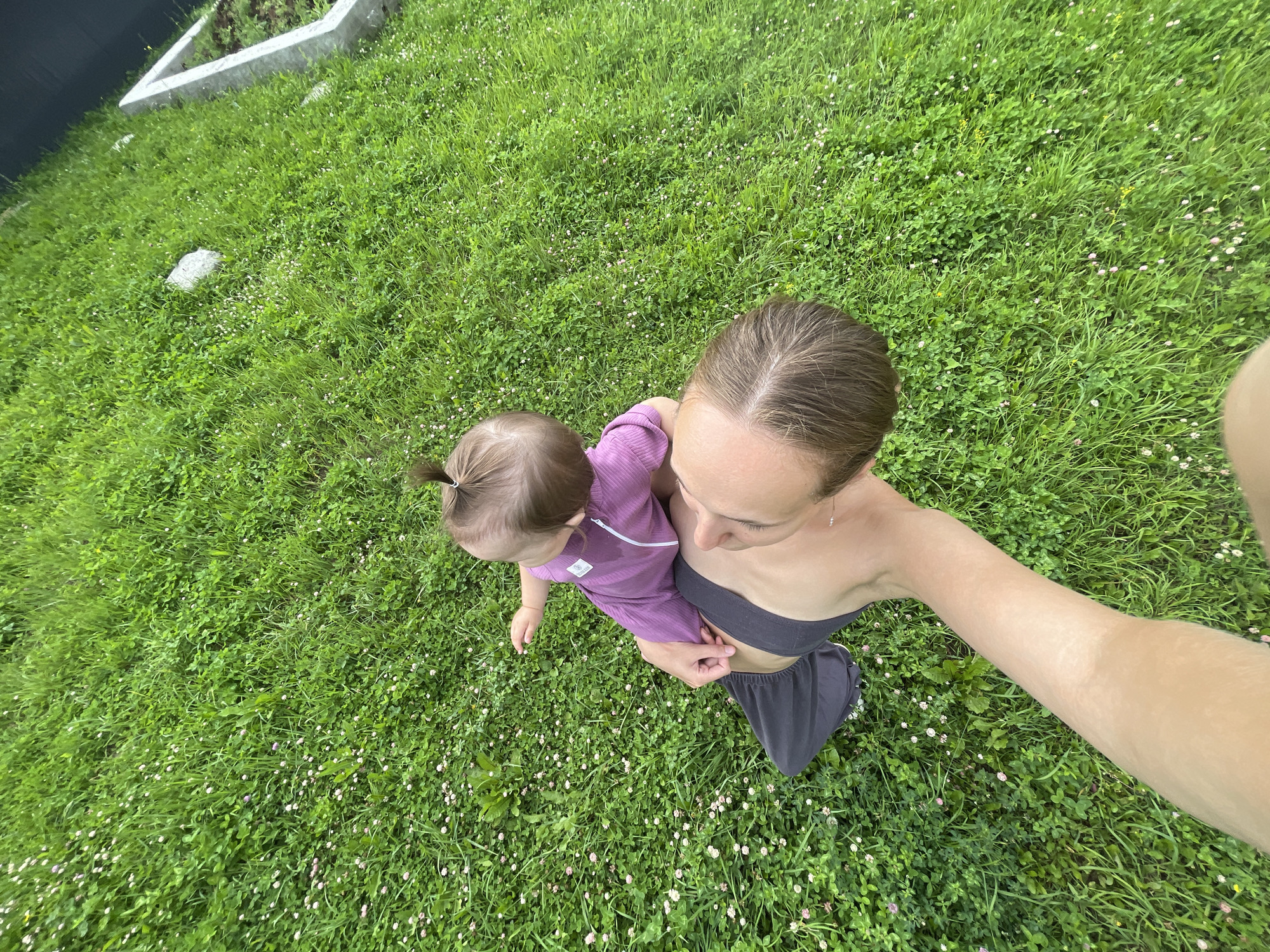 An overhead shot captures a person holding a young child in a grassy yard in the village of Podshivalovo, Russia. The person is wearing dark grey shorts and a black tank top, with their arm outstretched towards the camera. The child, dressed in a purple jumpsuit, is seated on the person's shoulders, facing away from the camera. The yard is covered in lush green grass, dotted with small white and pink clover flowers. In the background, a portion of a dark structure and a white concrete planter are visible. The sun appears to be shining, casting a natural light on the scene.