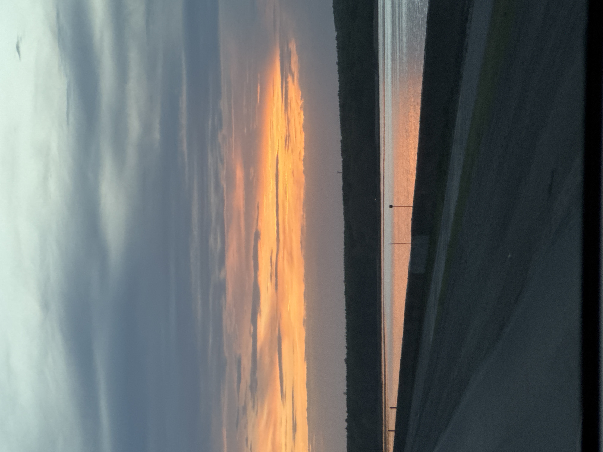 The image depicts a sunset over a body of water, likely a reservoir or large lake. The sky shows a gradient from blue/gray clouds to orange hues near the horizon, where the sun is likely setting. A strip of land with vegetation (trees or bushes) separates the sky from the water. The water reflects the colors of the sunset, creating a shimmering effect. There are what appear to be utility poles visible along the shoreline. The image appears to have been taken from a vehicle, as part of the interior is visible.
Given the context of Chaikovsky, Russia, it is highly probable that the body of water is the Votkinsk Reservoir, which is a major feature of the landscape in that region.