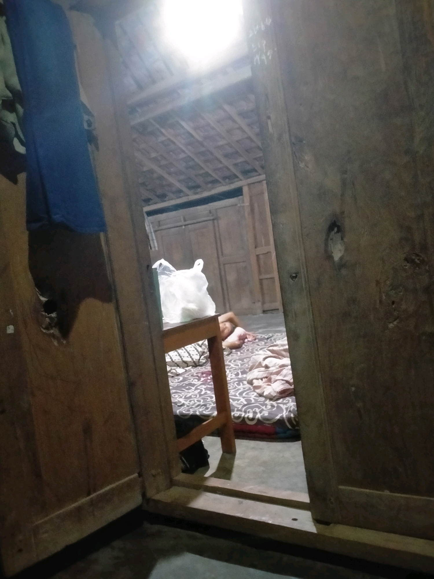 The image depicts an interior scene, possibly a room within a traditional house, viewed through an open doorway. The overall lighting is dim, with a bright light source emanating from the ceiling, creating a glare in the upper portion of the frame. The foreground is dominated by dark wooden structures, likely door frames and walls, with one wall showing a rough, unfinished texture and a small hole.
To the left, a blue garment hangs from a clothesline or hook. A large, white plastic bag is placed on a simple wooden table. In the background, a person, appearing to be a young female, lies on a mattress on the floor. She is seen from the side, with her body partially obscured by blankets and pillows. The mattress has a patterned cover, and the floor beneath it appears to be concrete or packed earth. Behind the person, there are wooden doors and walls, suggesting the interior of a dwelling. The ceiling structure, visible through the doorway, appears to be made of wooden beams and tiles, characteristic of traditional Indonesian architecture.
There are white markings on the wooden door frame on the right side of the image, which appear to be handwritten characters or numbers. One crop shows these markings more clearly, appearing to be "2014" followed by other illegible characters.
The context provided indicates this scene is located in Playen, Indonesia. This implies a setting typical of rural or semi-rural Indonesian housing, characterized by natural materials and a simpler construction style. The image captures a candid moment, potentially suggesting a domestic or personal space.